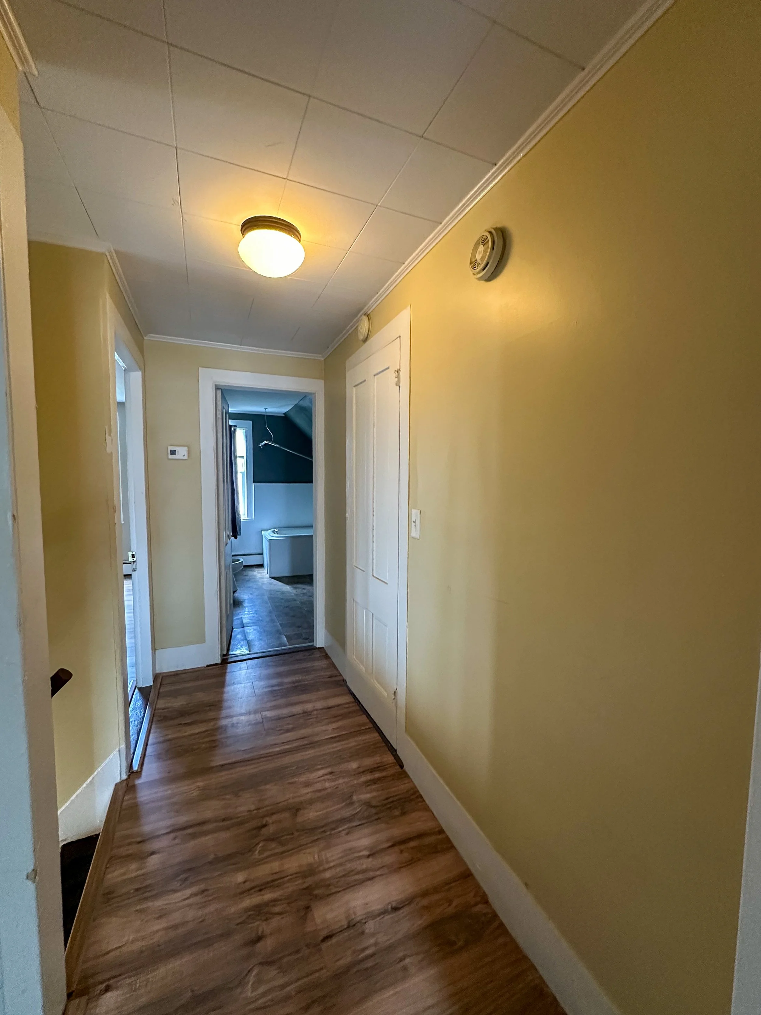 A hallway with yellow painted walls, wooden hardwood floor, a white ceiling with flush mount ceiling light, and smoke detectors on the wall. At the end of the hallway, there is a door leading to a bathroom with blue walls and tile flooring.