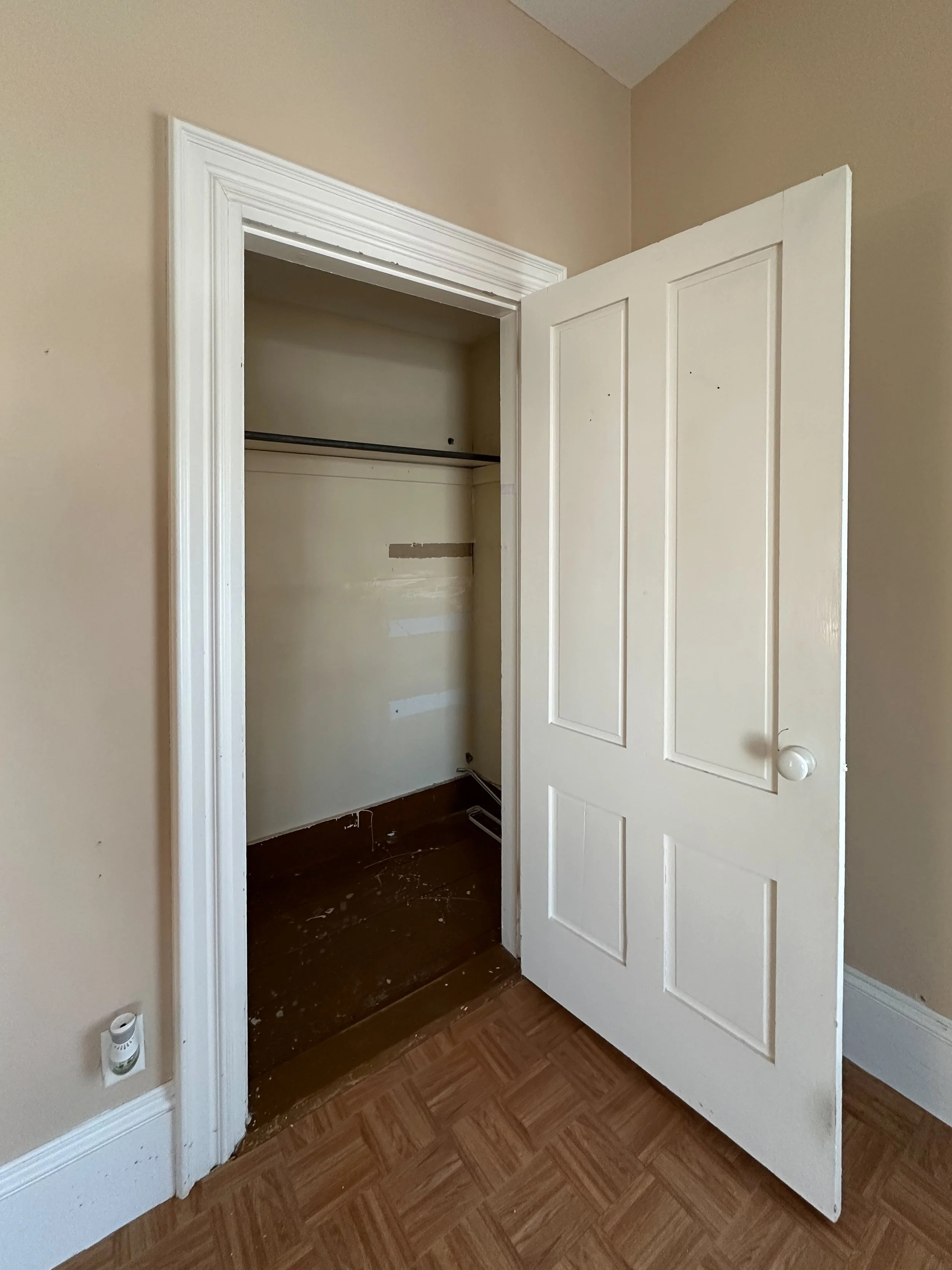 Empty closet with an open door in a room with beige walls and hardwood floors.