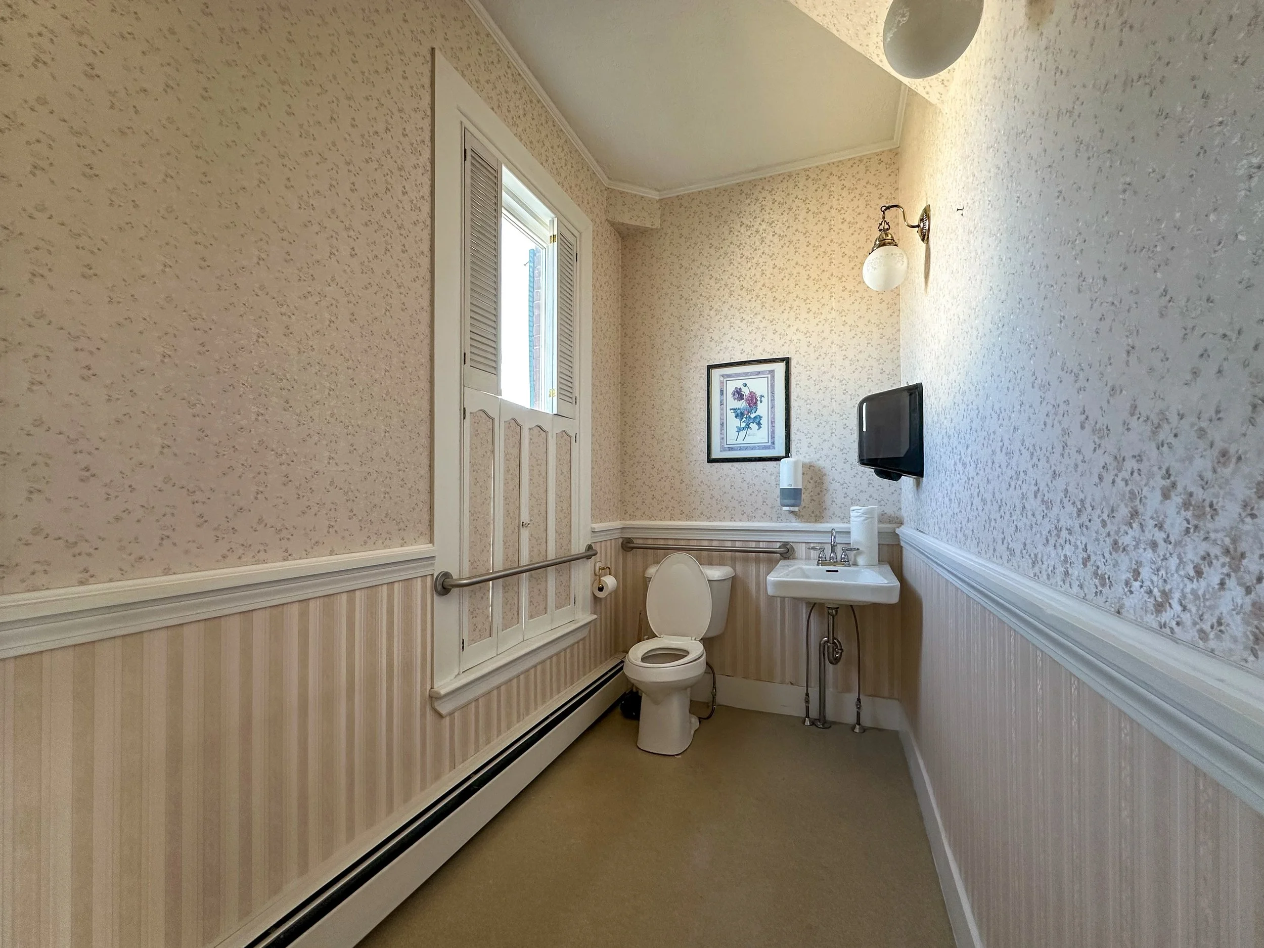 A bathroom with floral wallpaper, a window with louvered shutters, a picture frame on the wall, a wall-mounted light fixture, a toilet, a small sink with a paper towel dispenser nearby, and a bath safety grab bar.