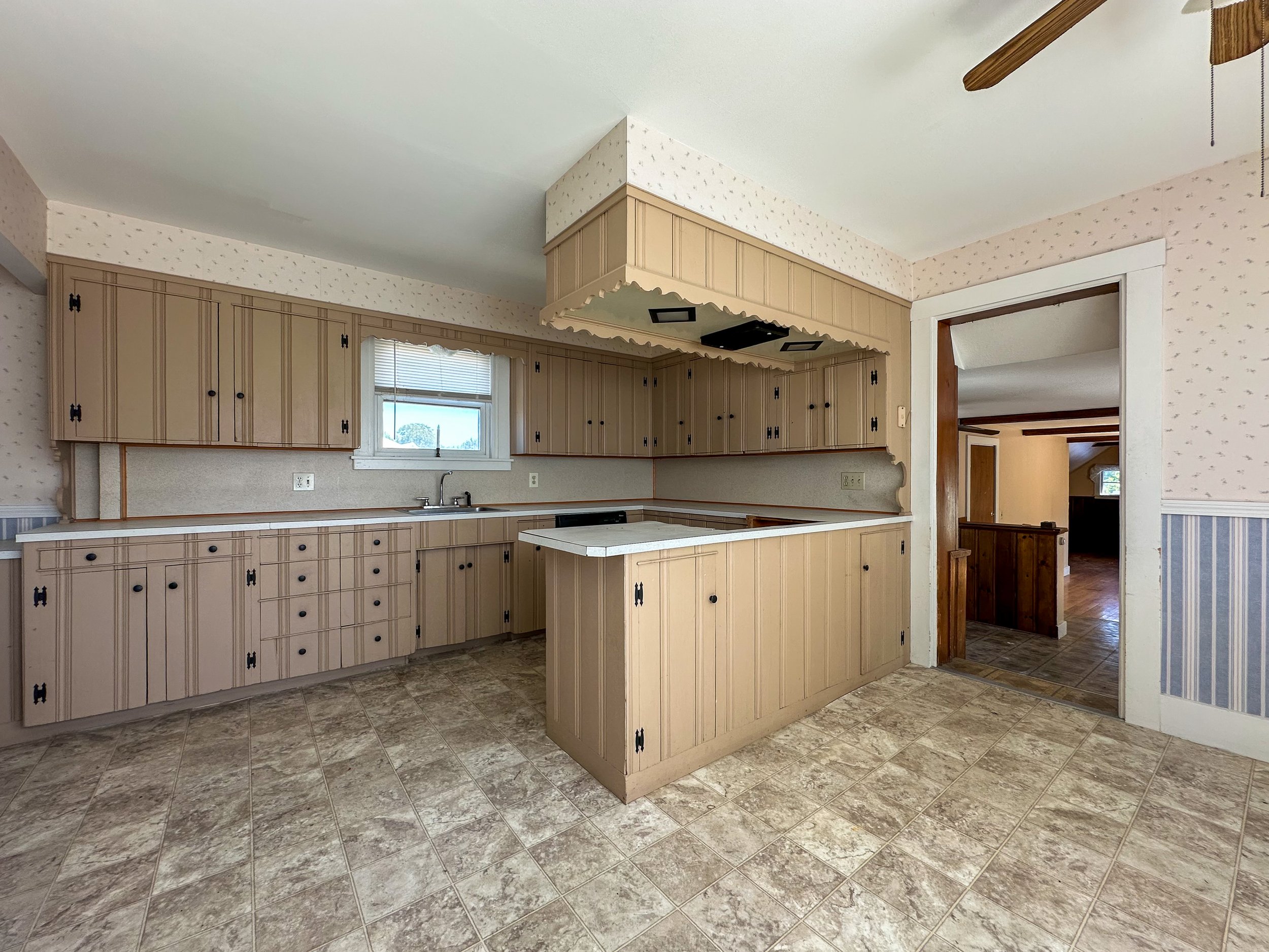 A kitchen with beige cabinets, a small window, a countertop with a sink, and a large kitchen island with a matching beige countertop. The floor is tiled with a beige and brown pattern. There is a doorway leading to another room and a ceiling fan with