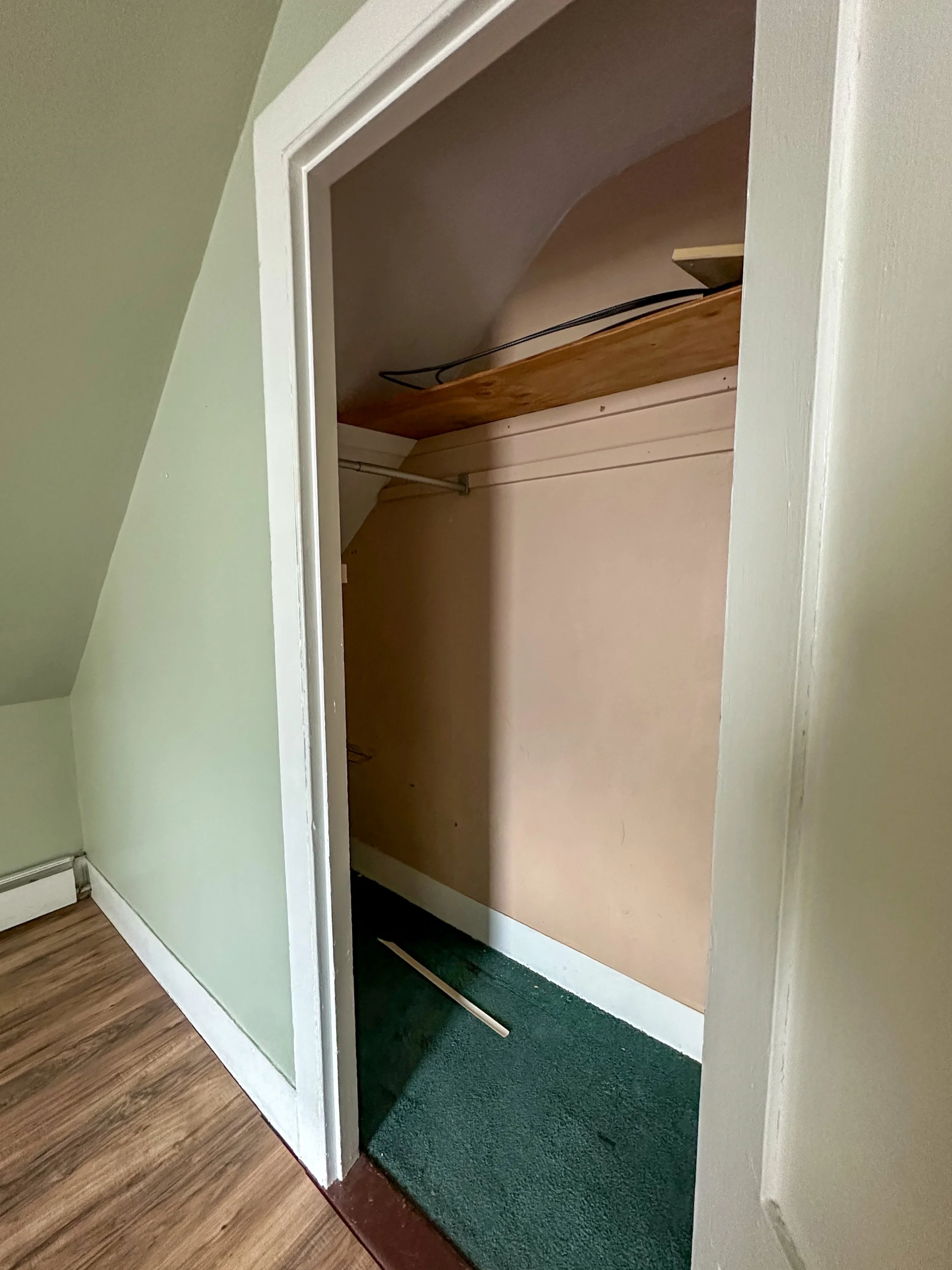 Empty closet with a wooden shelf and green carpet, partially open door, beige walls, and wooden floor.