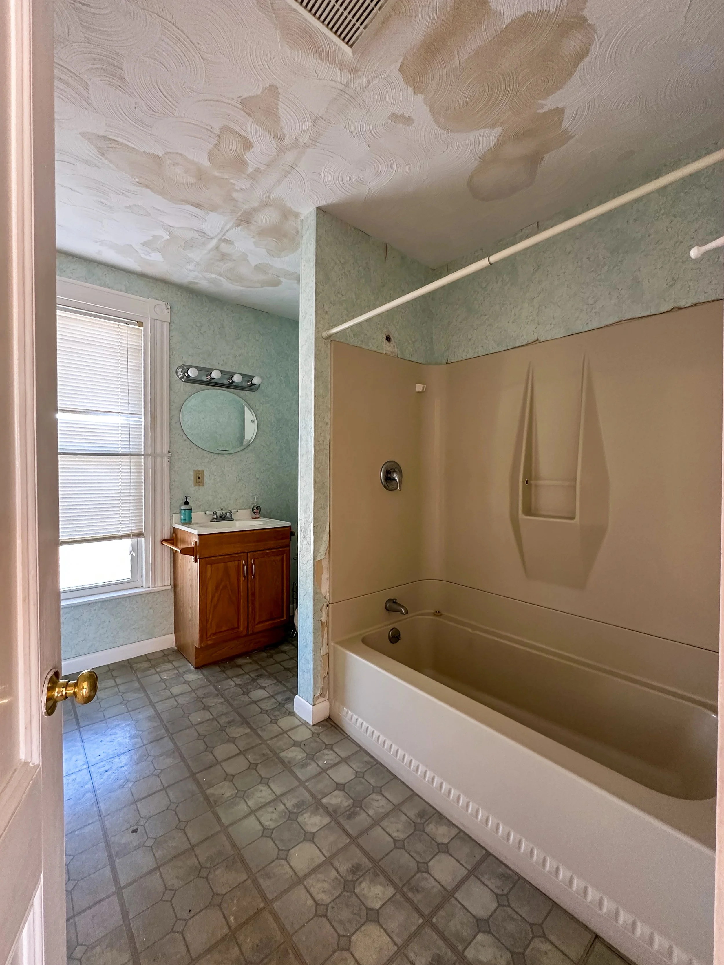 A bathroom with a bathtub, shower head, sink, mirror, wooden cabinet, window with blinds, and tiled floor. The ceiling has water stains or damage.
