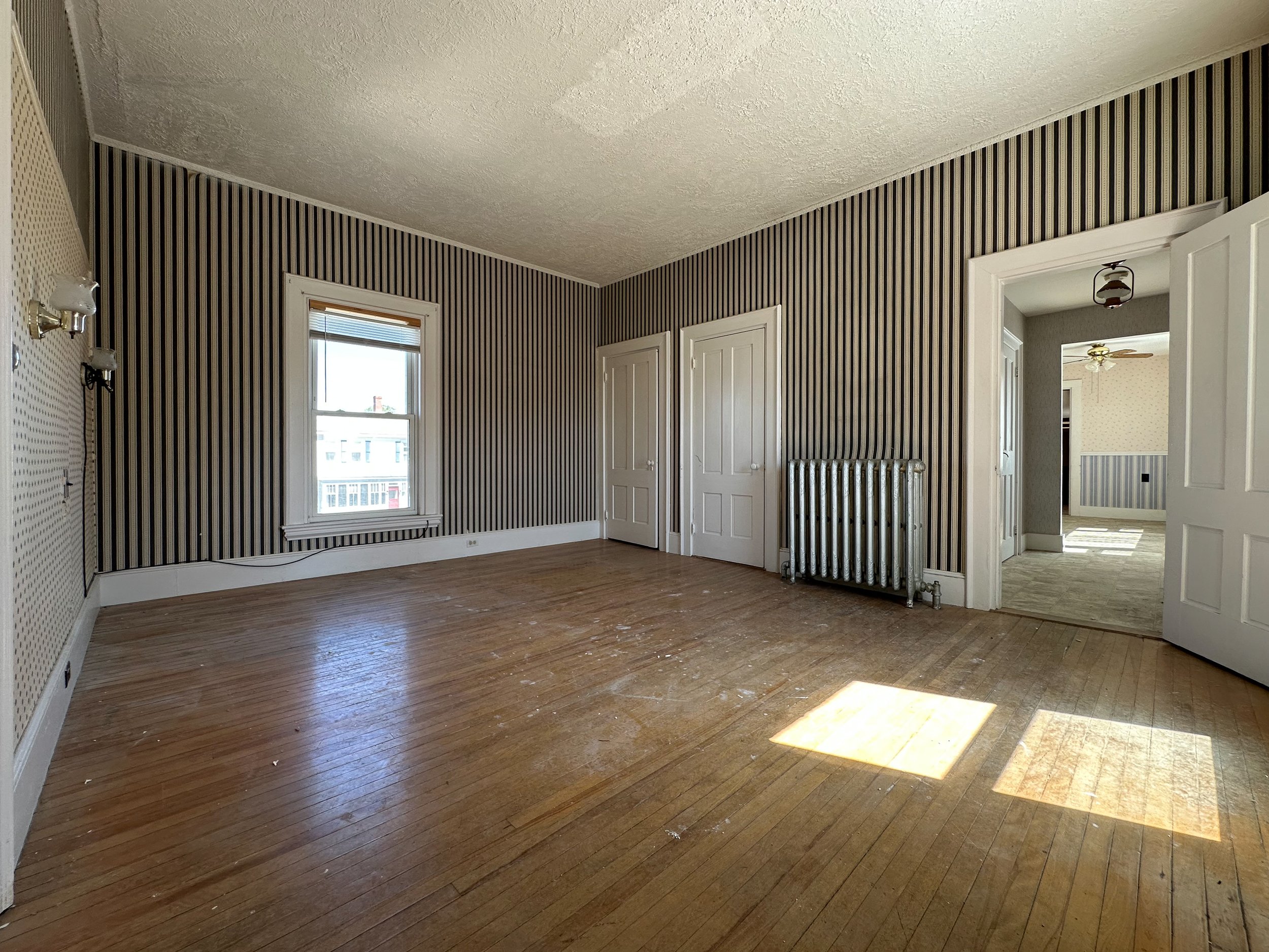 Empty room with hardwood floor, striped wallpaper, a window with blinds, a radiator, and an open doorway leading to another room with similar decor and a ceiling fan.