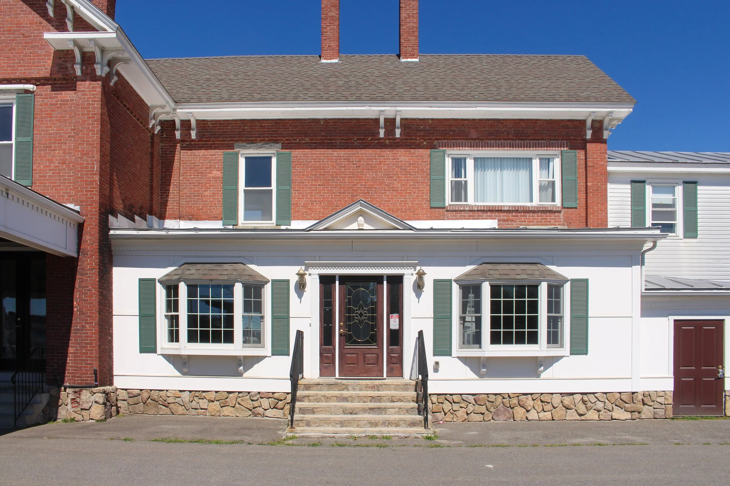 Front view of a multi-story brick house with large windows, green shutters, and stone steps leading to a wooden door. The house has a detailed roof with two chimneys and white trim. The sky is clear and blue.
