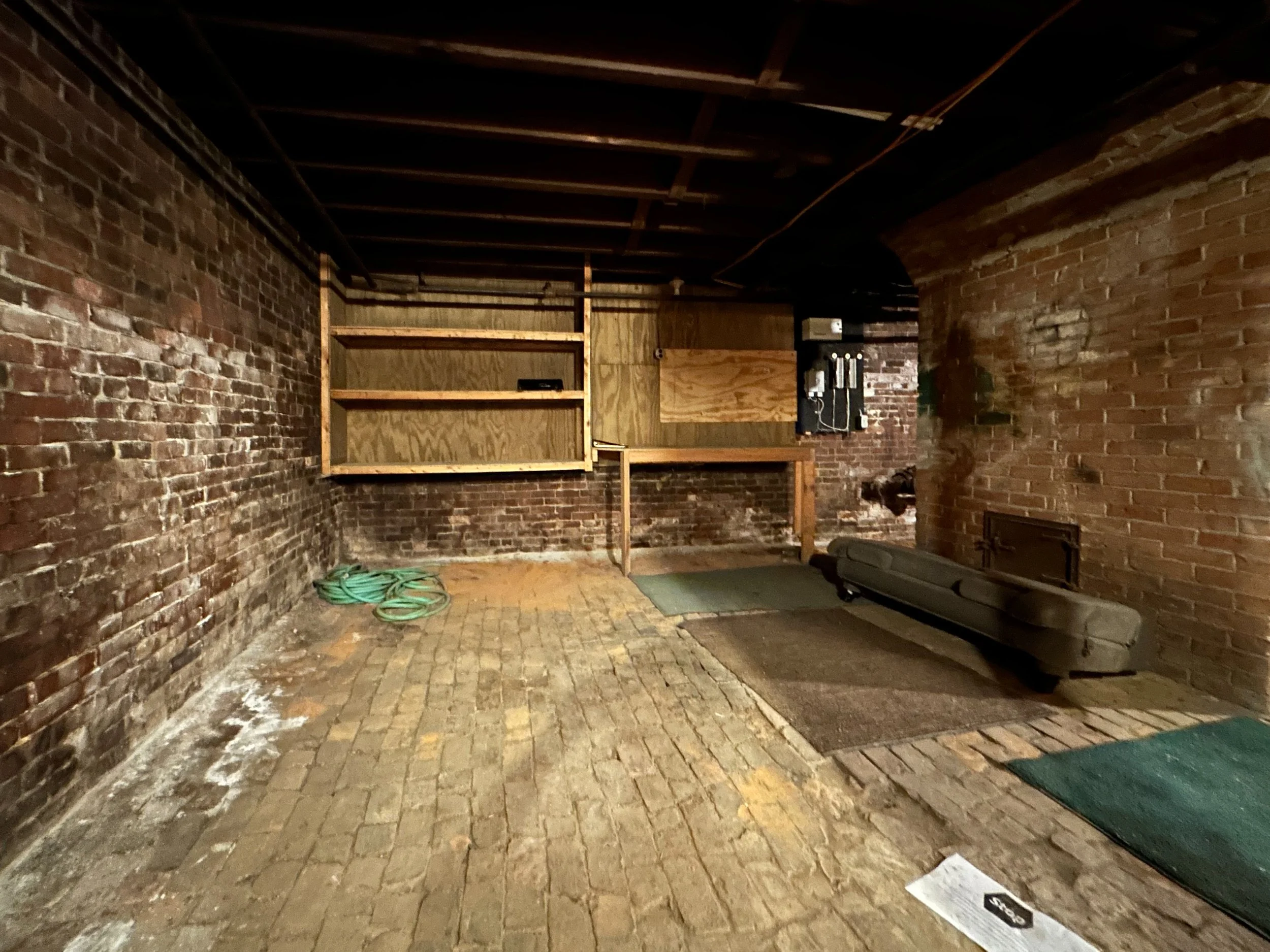 This image shows an unfinished basement with brick walls and a brick floor. There is a wooden structure on one side, some electrical wiring and a circuit box on the wall, and a few shelves. There is a green hose on the floor and a treadmill in the co