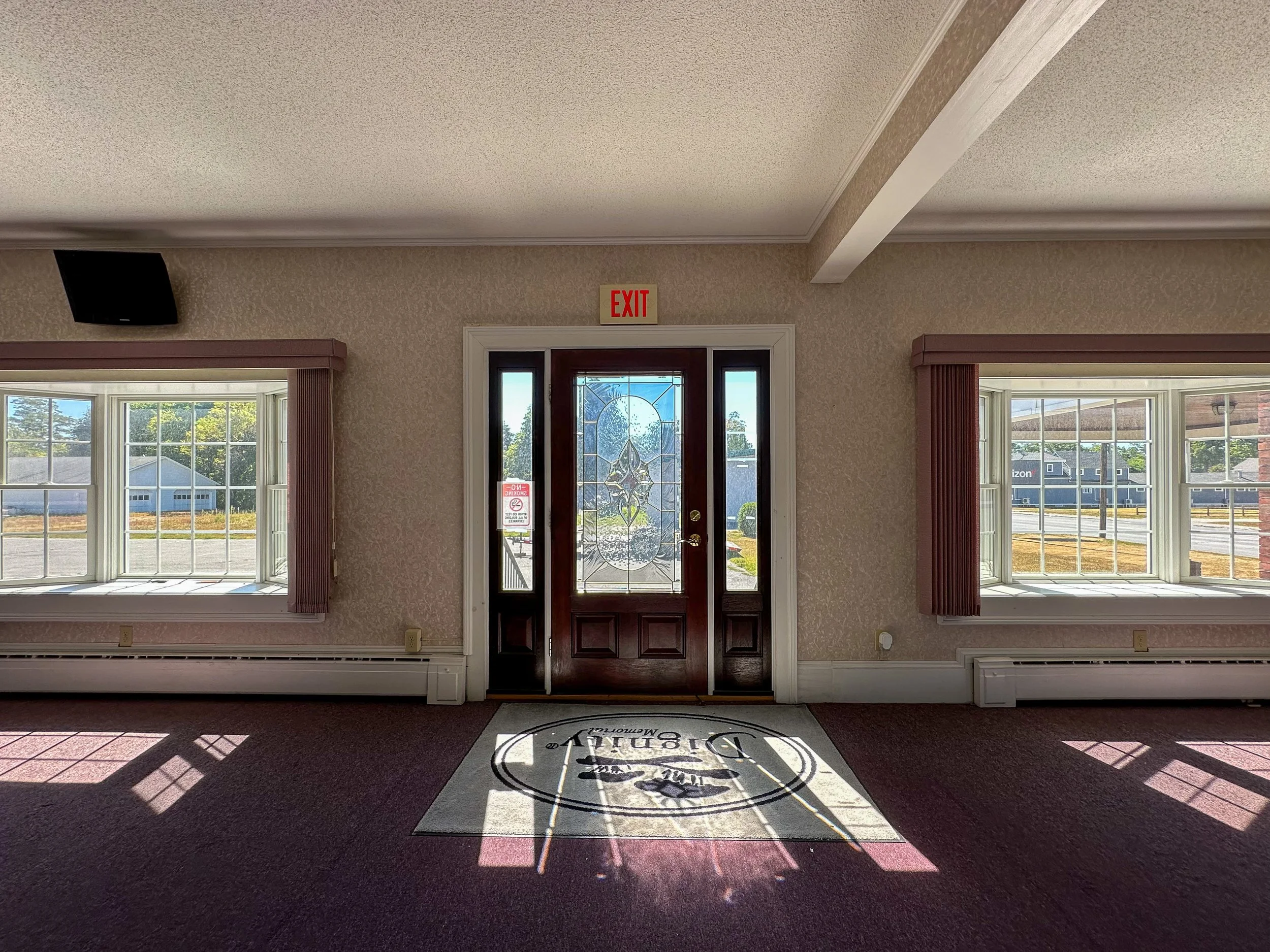 A view of the interior of a house near the entrance with sunlight streaming through the windows, casting shadows on the carpeted floor with a welcome mat.