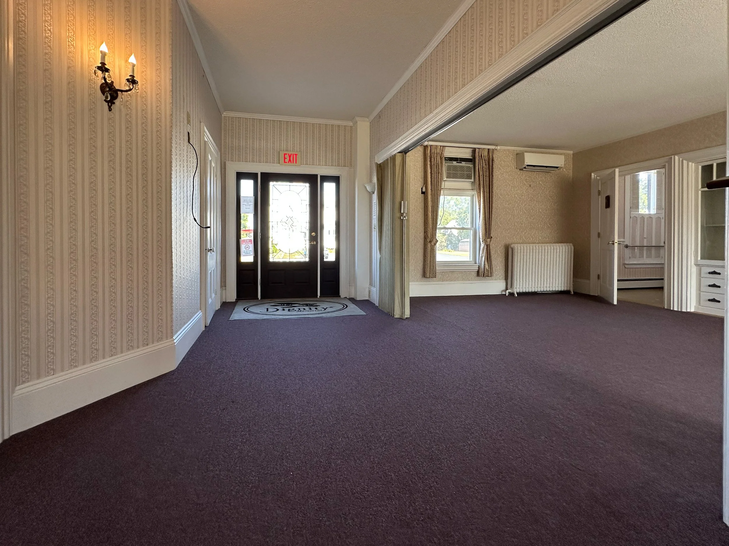 Interior view of an entryway with a black front door, a welcome mat, beige wallpaper, pink curtains, a radiator, and windows allowing natural light.