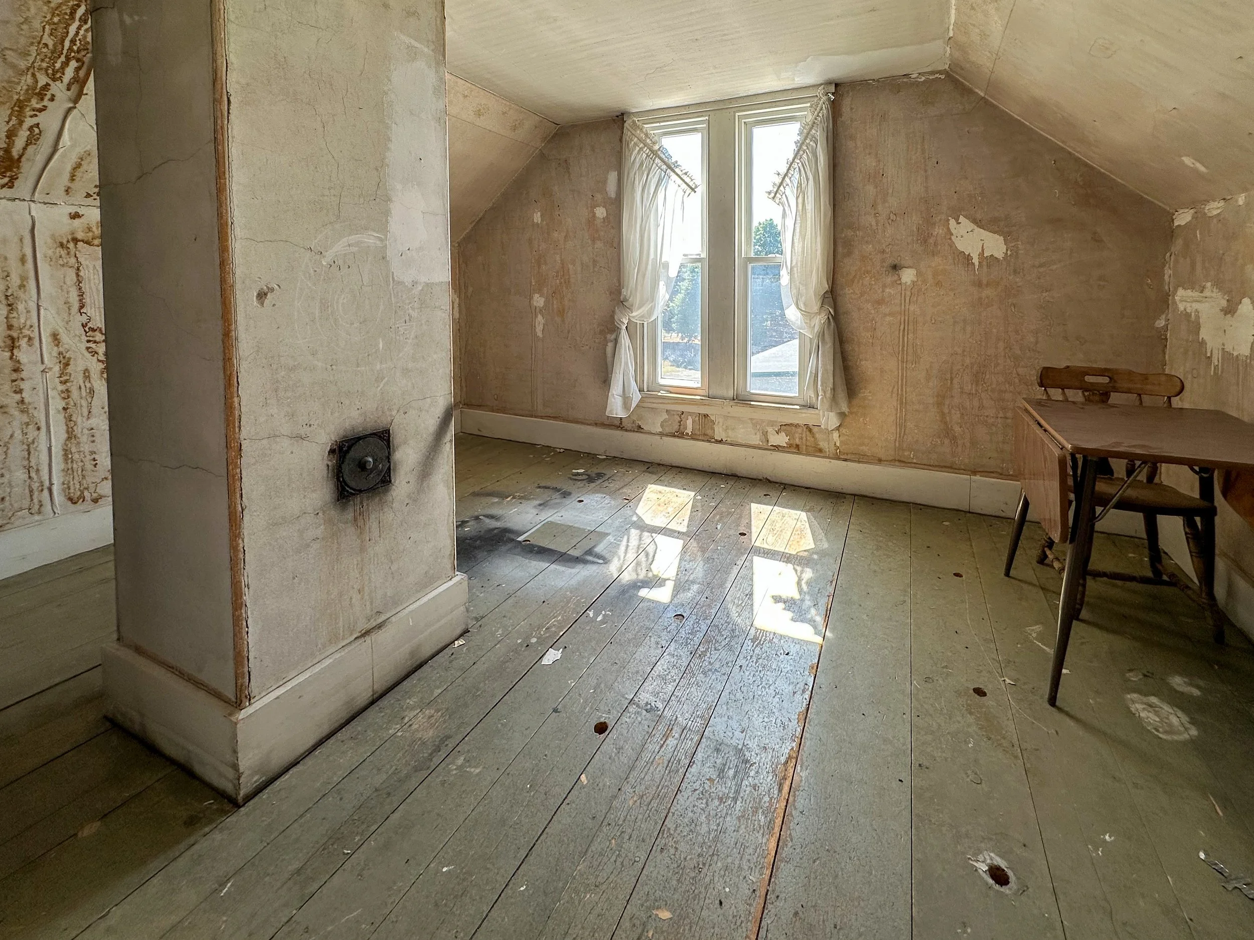 An empty, worn room with wooden floorboards, peeling wallpaper, and a window with white curtains, with sunlight shining through.