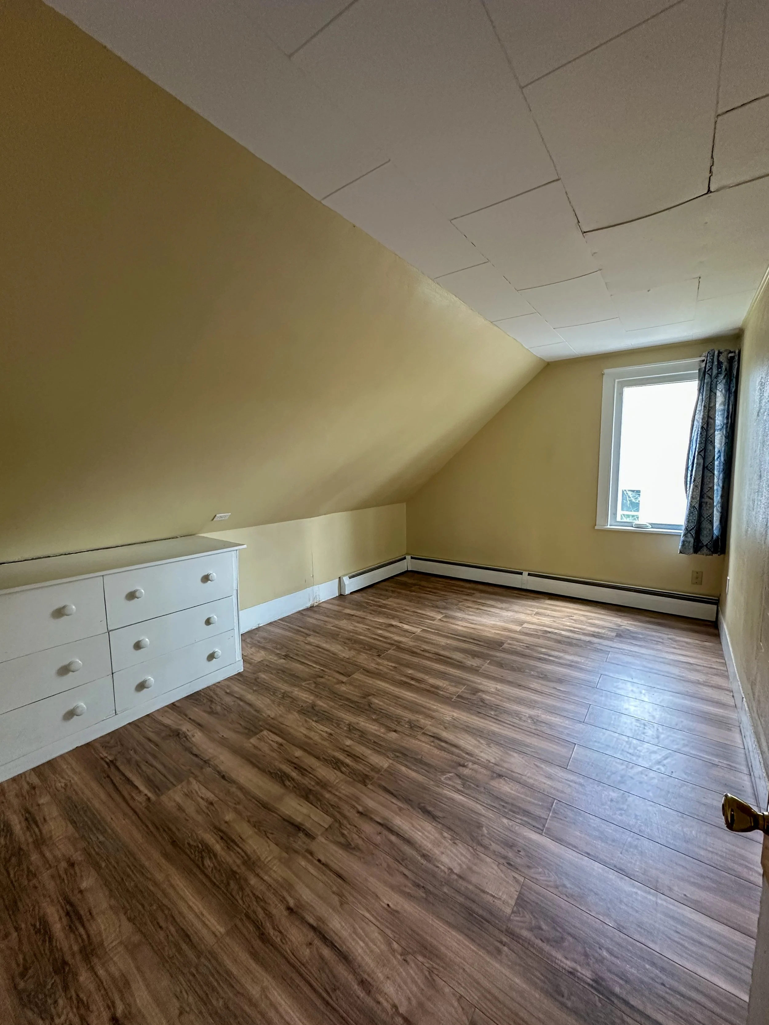 An empty room with yellow walls, wood flooring, a window with a dark patterned curtain, a white dresser, and a sloped ceiling with visible joints in the drywall.