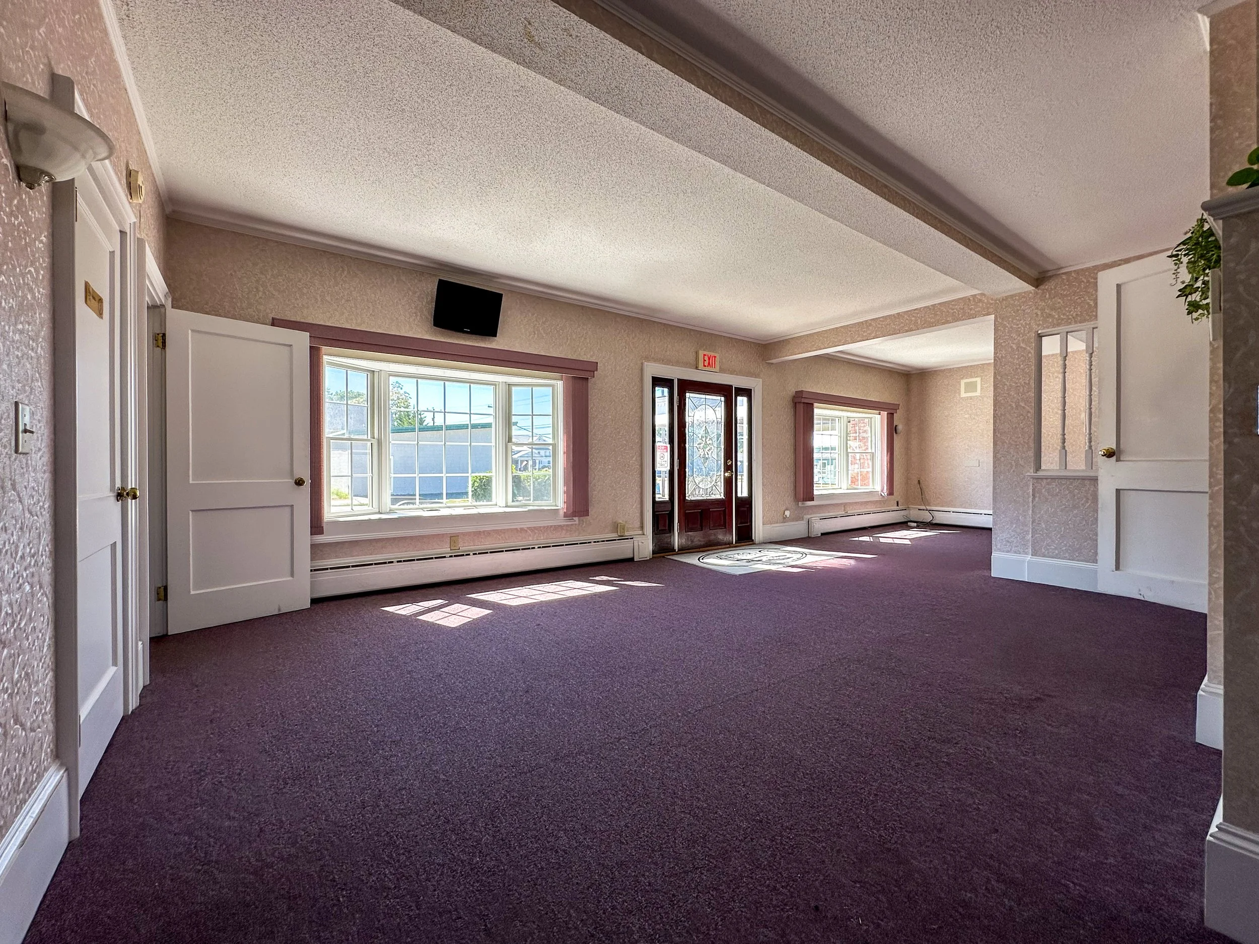 Interior of a house with white walls, large windows allowing natural light, an entry door with decorative glass, and a brown carpeted floor.