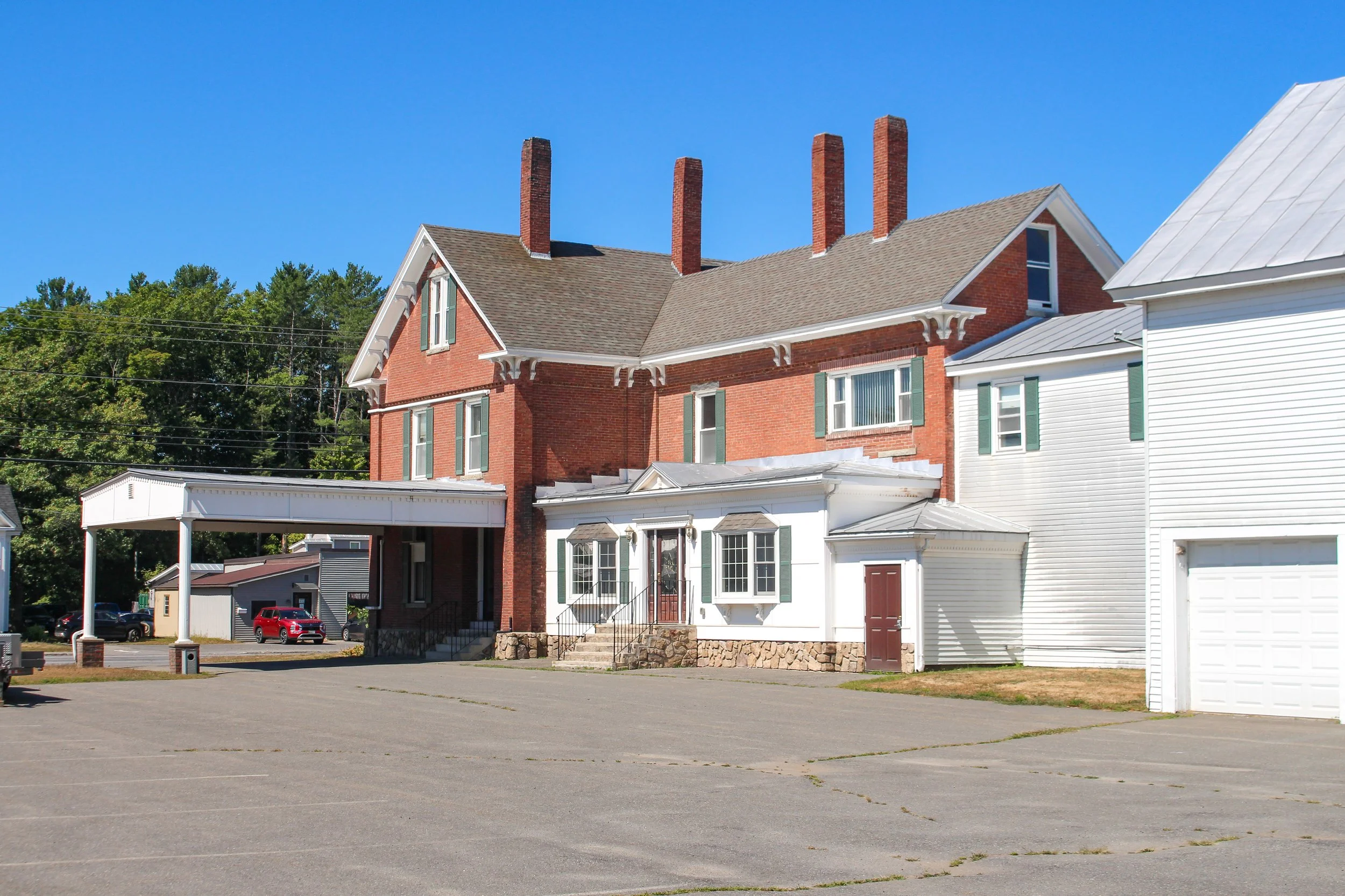A large brick house with multiple chimneys and green shutters on the windows. The house has a front porch with steps leading up to it, and a white garage is attached to the house. There is a parking lot with several parked cars, and green trees and a