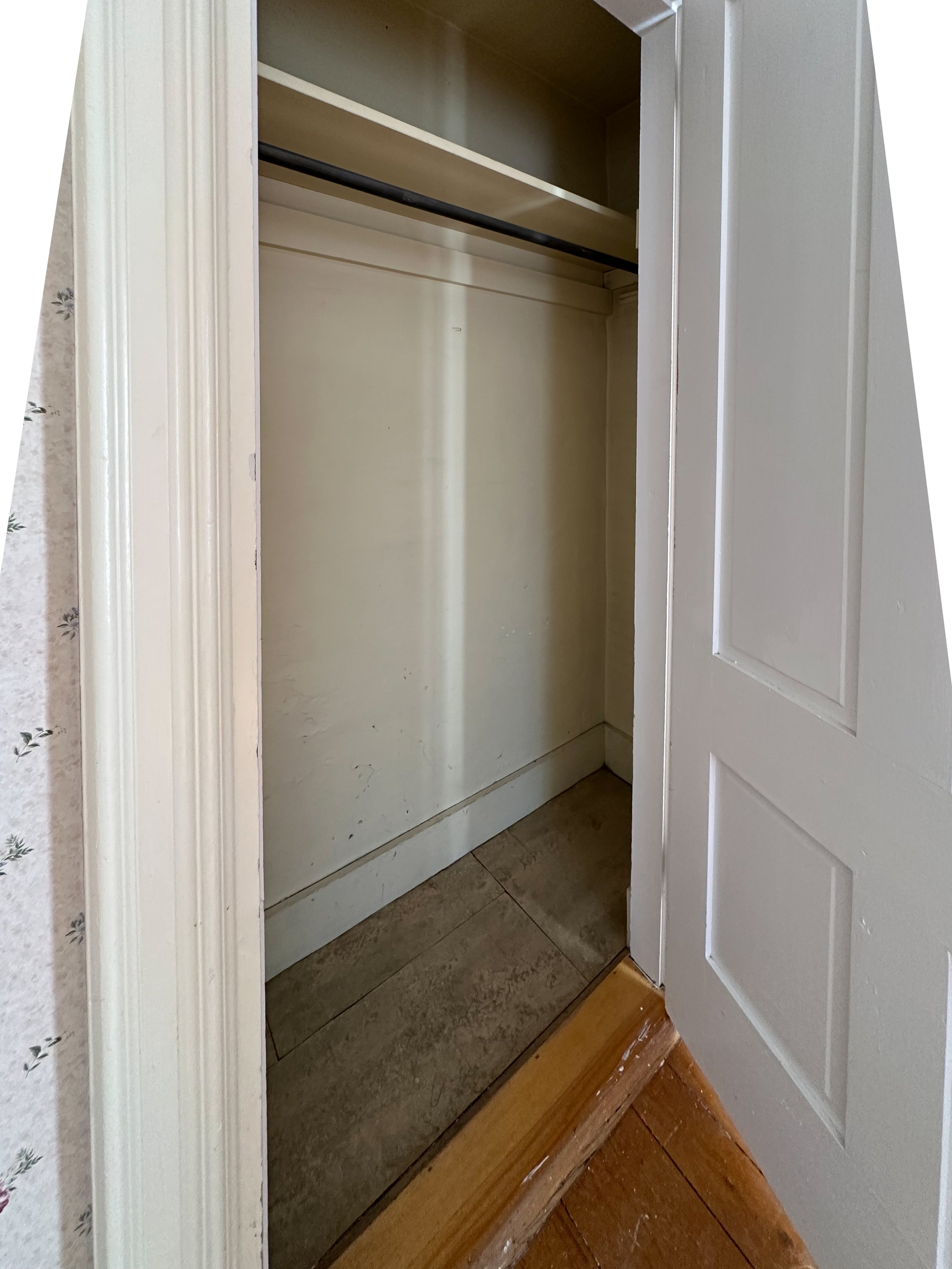 An empty closet with a hanging rod, inside a room with tiled flooring, with a partly open white door.