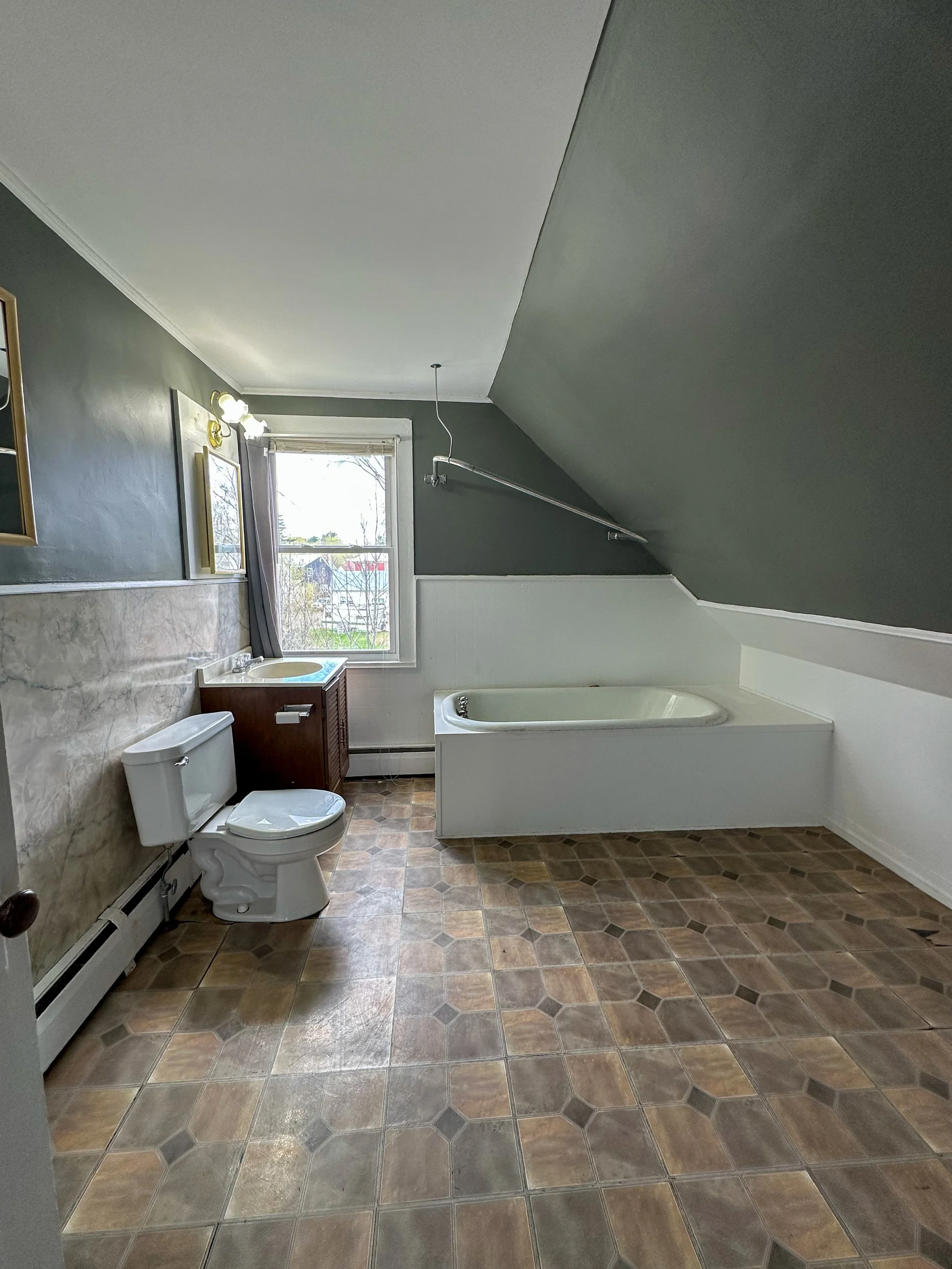 A bathroom with a toilet, a sink with a cabinet, and a bathtub. The floor has brown patterned tiles, and there is a window with gray curtains. The walls are painted dark green, and a mirror hangs above the sink. Ceiling lighting illuminates the room.