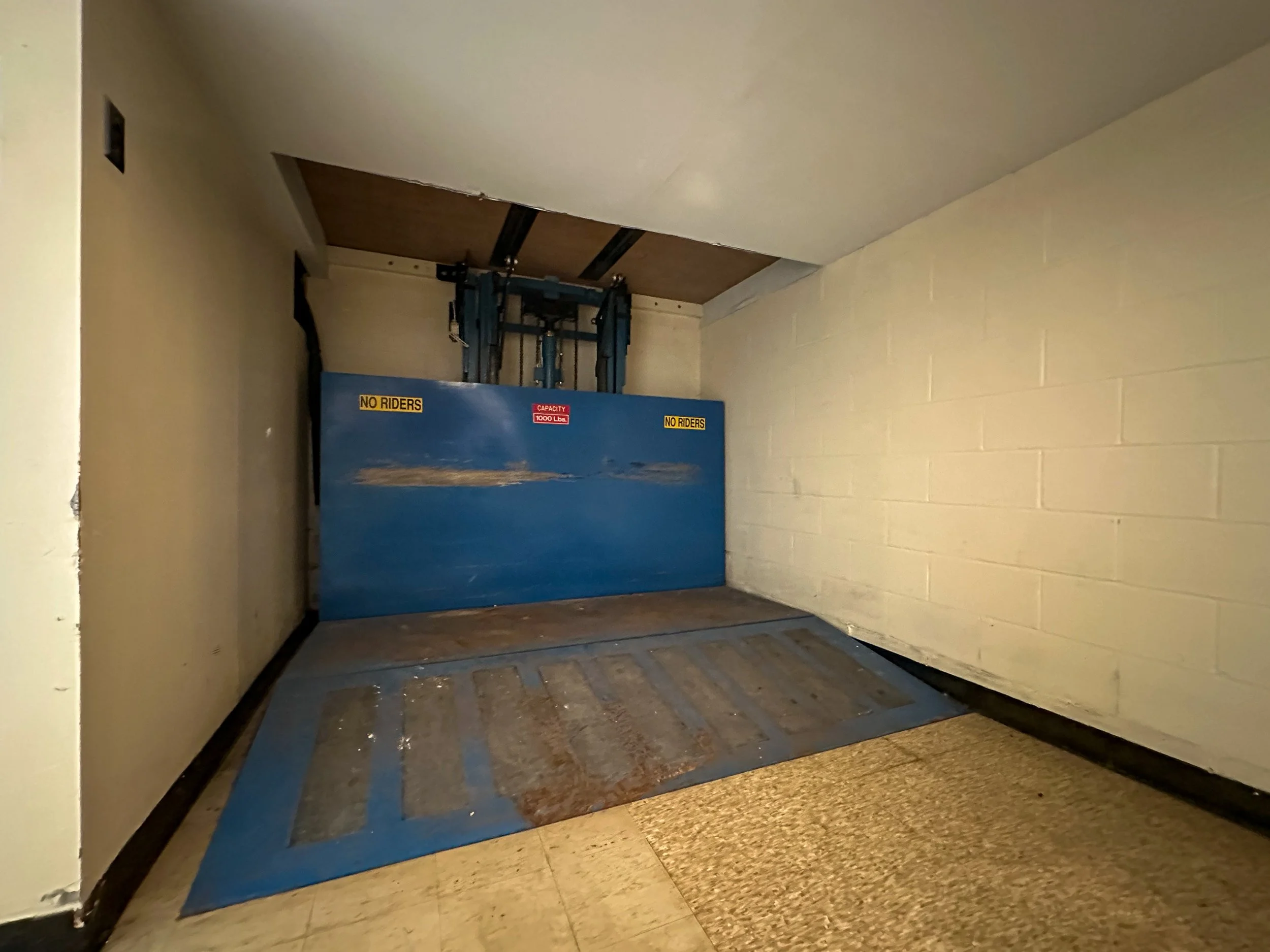A safety lift outside a storage room door with warning signs, positioned against a beige cinder block wall, on a tiled floor.