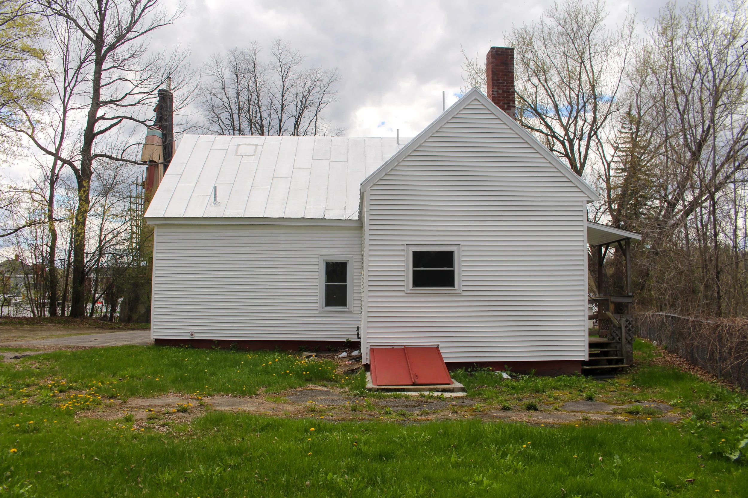 White house exterior with green lawn, trees in background, cloudy sky, red rooftop vent, chimney, and a staircase on the right side.