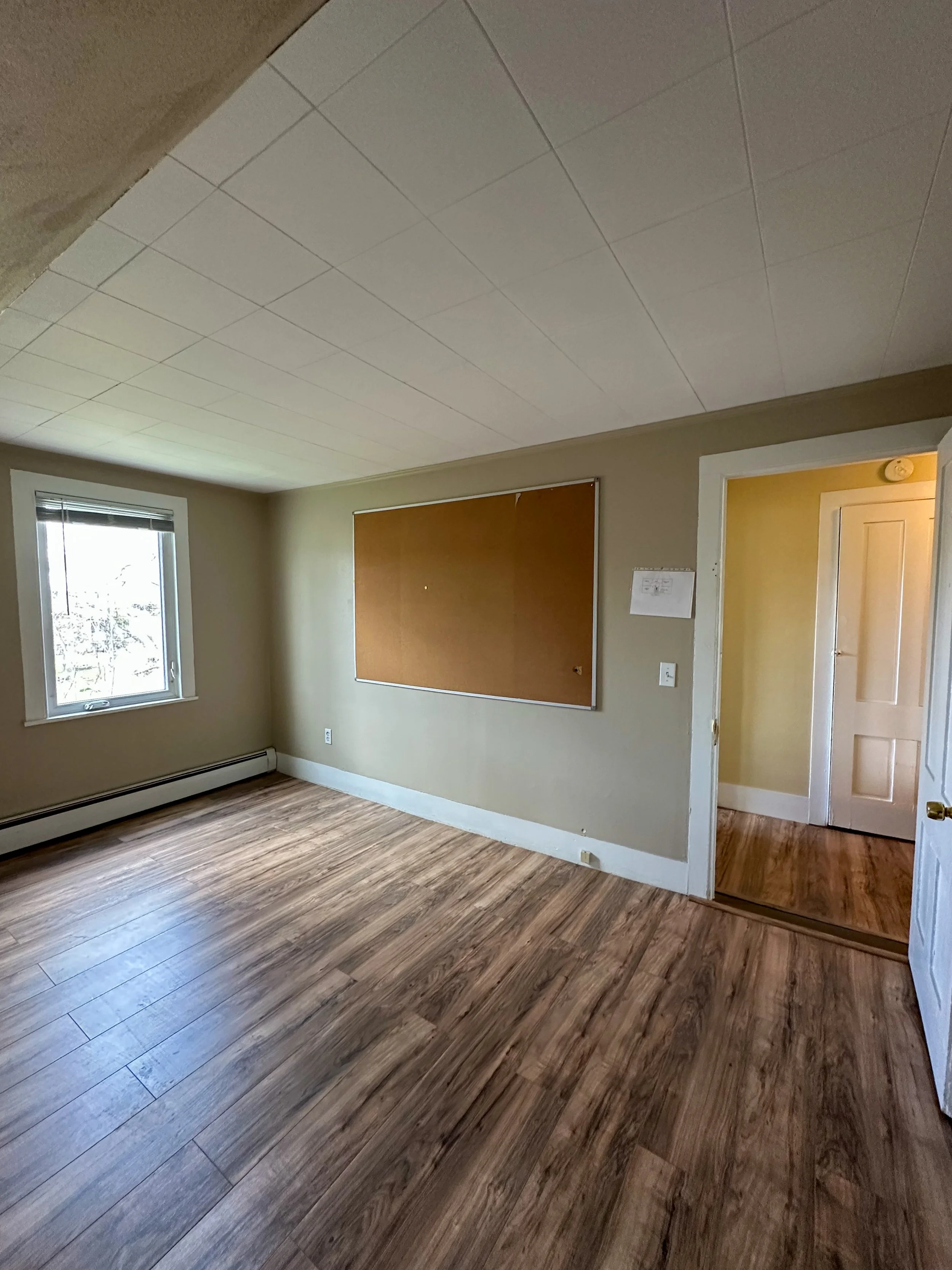 Empty room with hardwood floors, a window with blinds, a large corkboard on the wall, and an open door leading to another room with yellow walls.