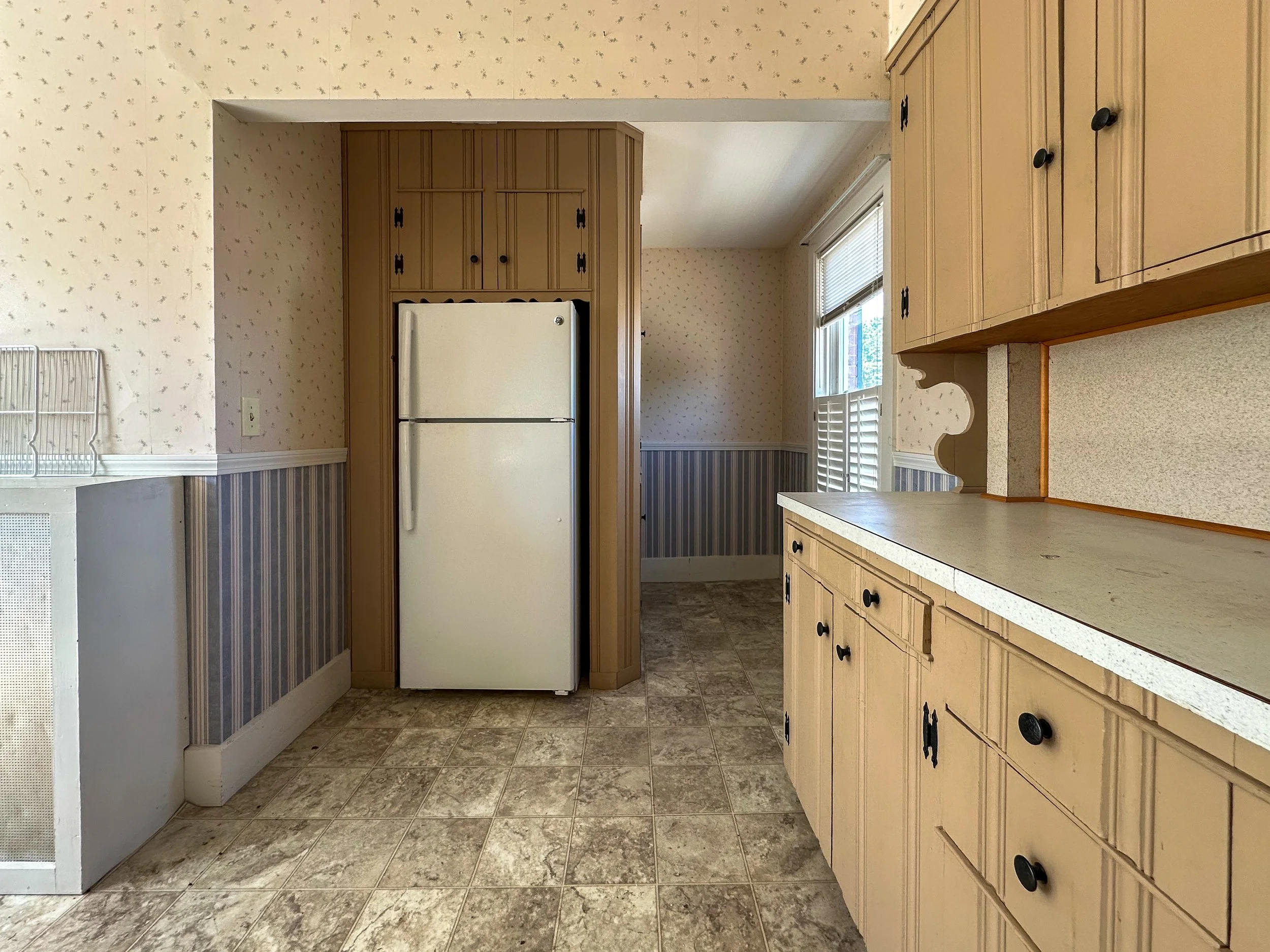A kitchen with beige cabinets, a white refrigerator, a window with blinds, and a countertop with no objects on it. The floor is tiled with a patterned design.