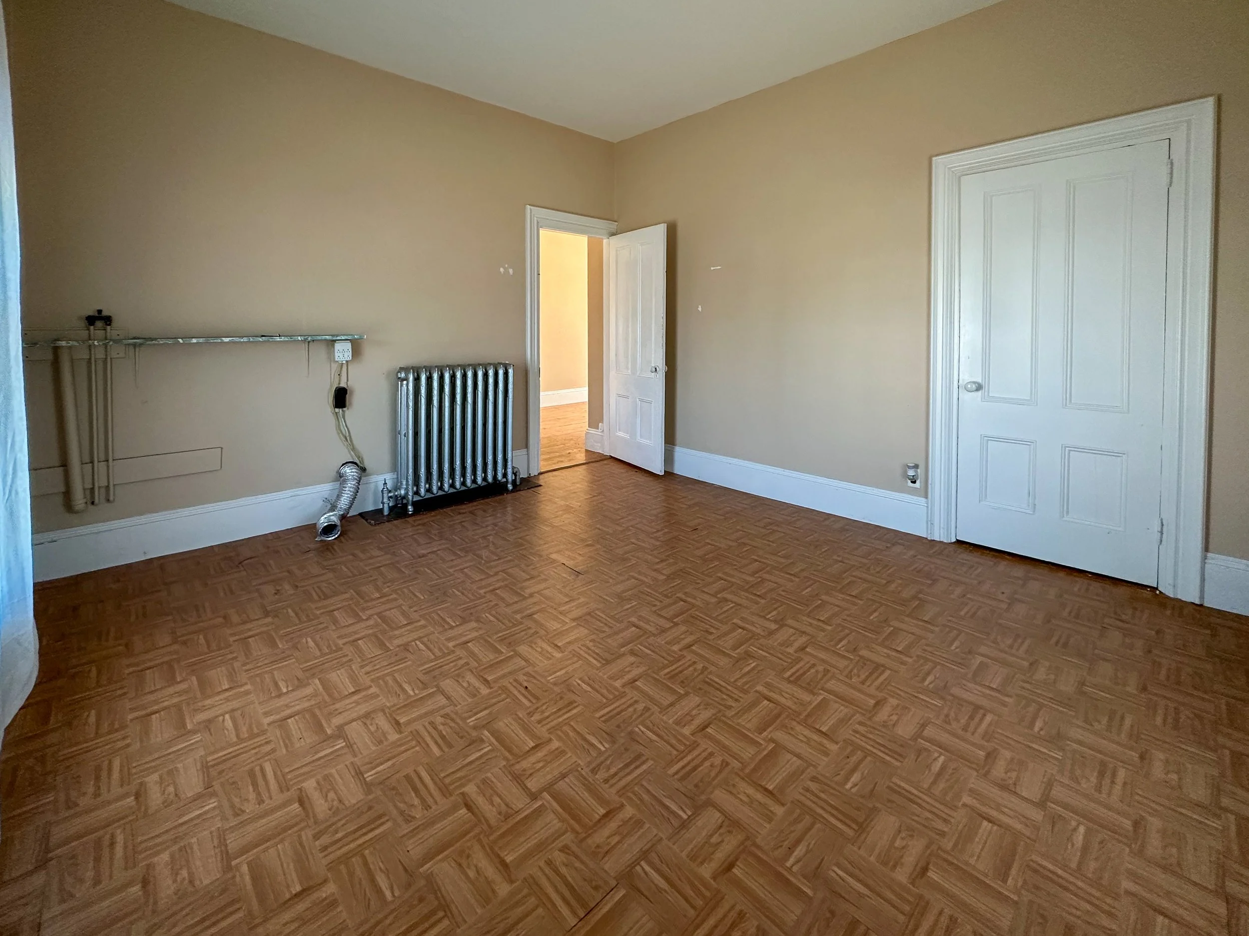 Empty room with beige walls, hardwood floor, radiator, and white paneled doors.