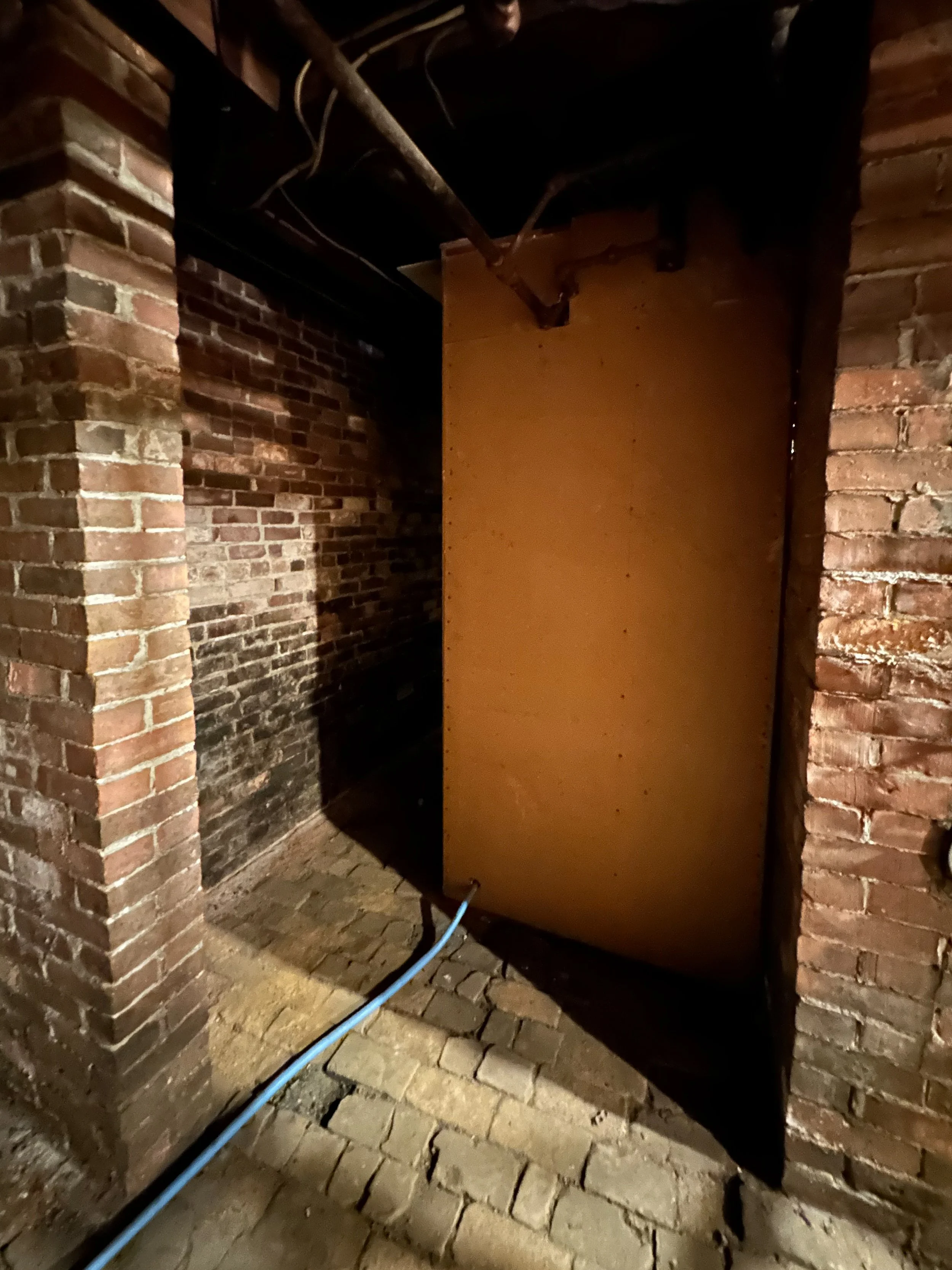 Brick basement with exposed pipes and a large wooden panel on a metal stand, with a blue conduit pipe running along the brick floor.