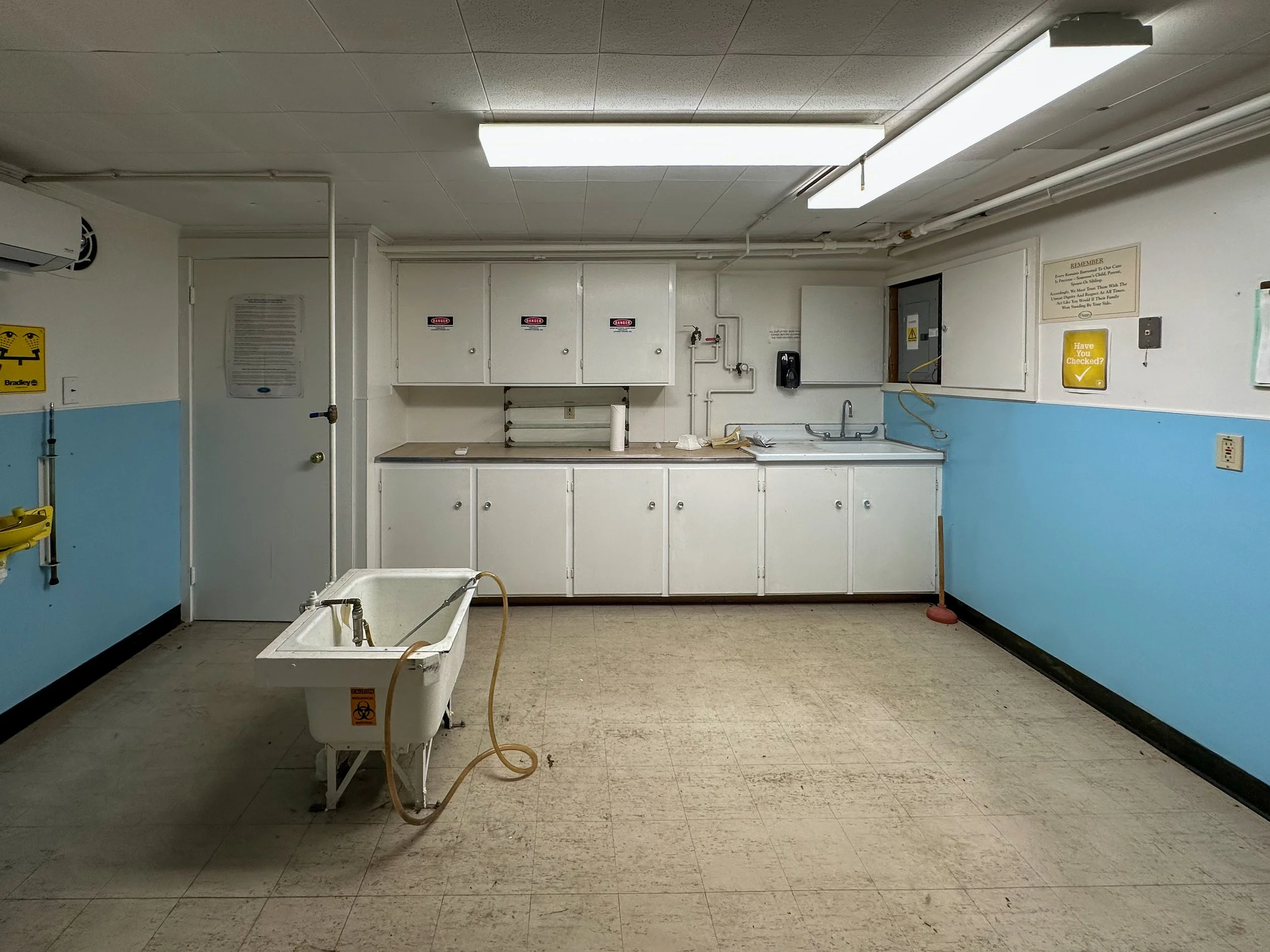 A sterile hospital room with a wall-mounted sink and cabinets, a raised bathtub with hoses attached, and warning signs on the walls and equipment.