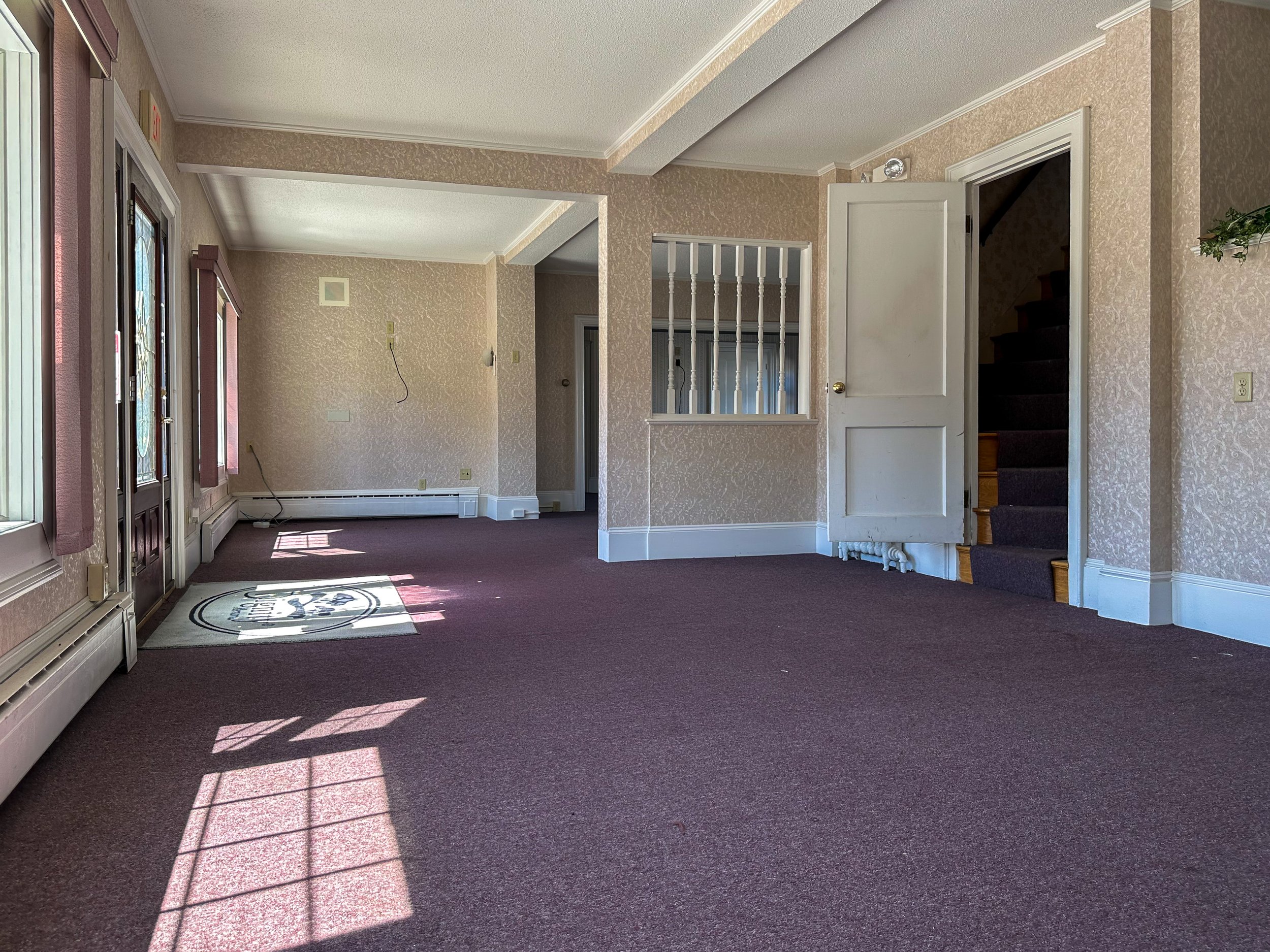 Interior of an empty room with pink patterned wallpaper, a purple carpet, and sunlight streaming through windows, creating shadows on the floor.