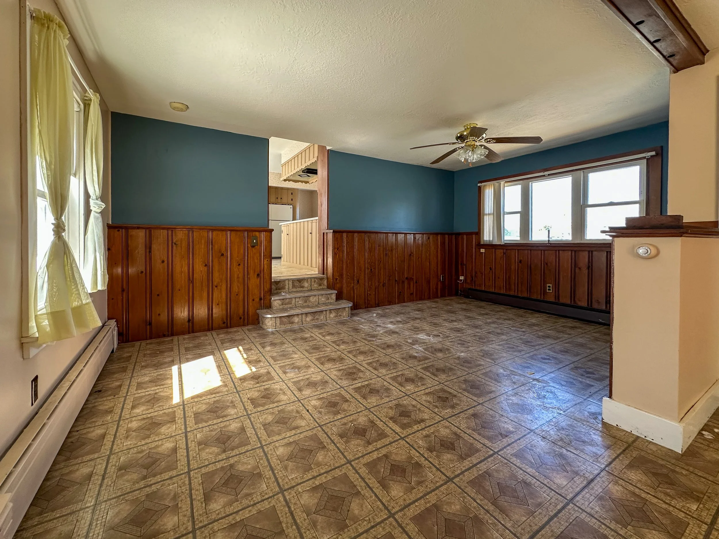 Living room with blue walls, wooden wainscoting, tiled floor, large windows with curtains, ceiling fan, and stairs leading to another level.