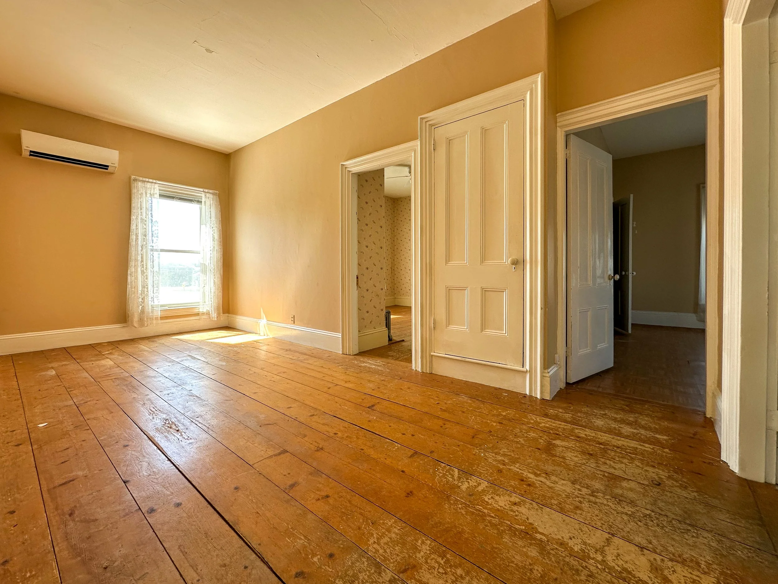 Empty room with yellow walls, hardwood floors, a window with sheer curtains, and multiple doorways leading to other rooms.