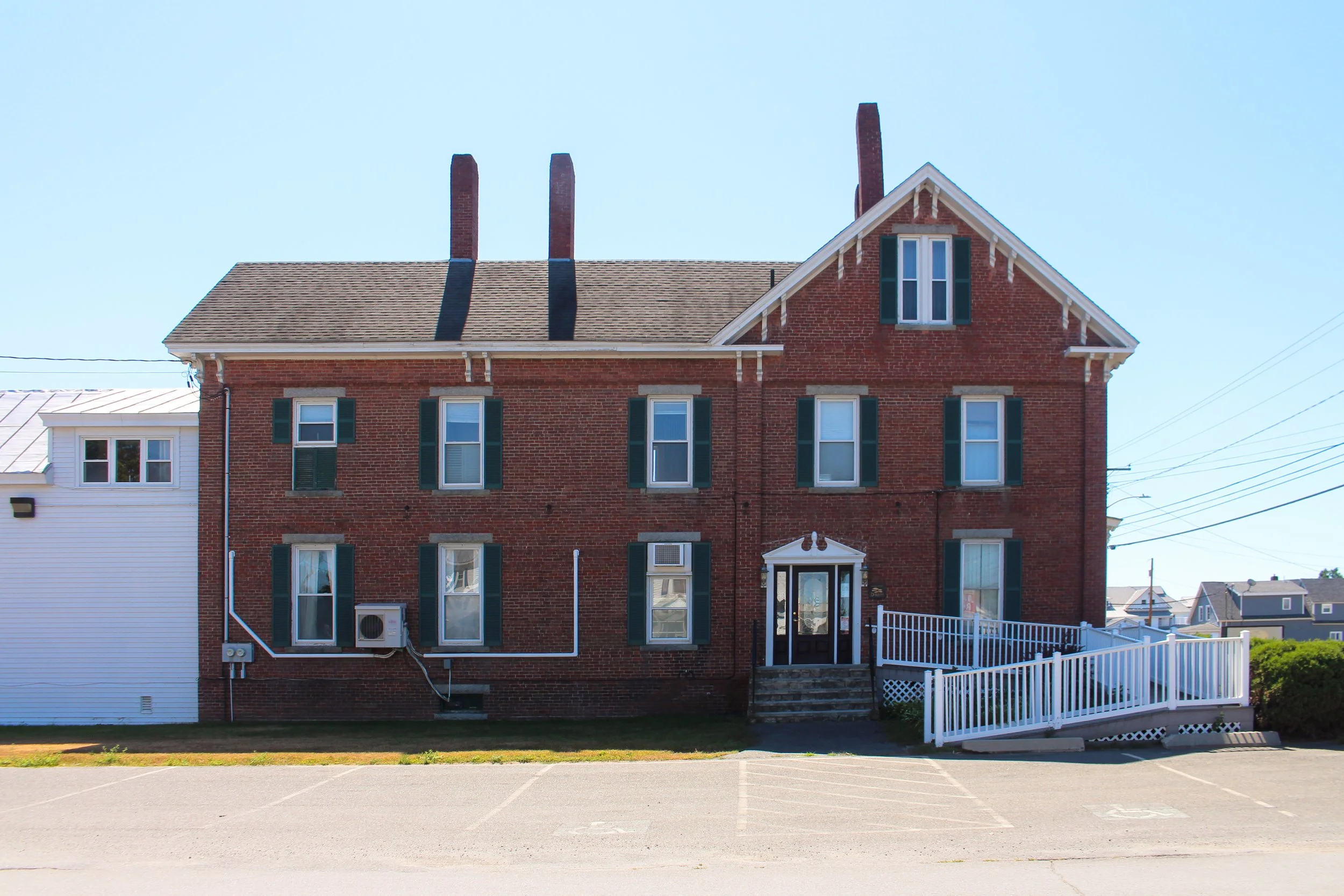 A multi-story brick building with green shutters and a white door, fenced with a white picket fence, under a clear blue sky.