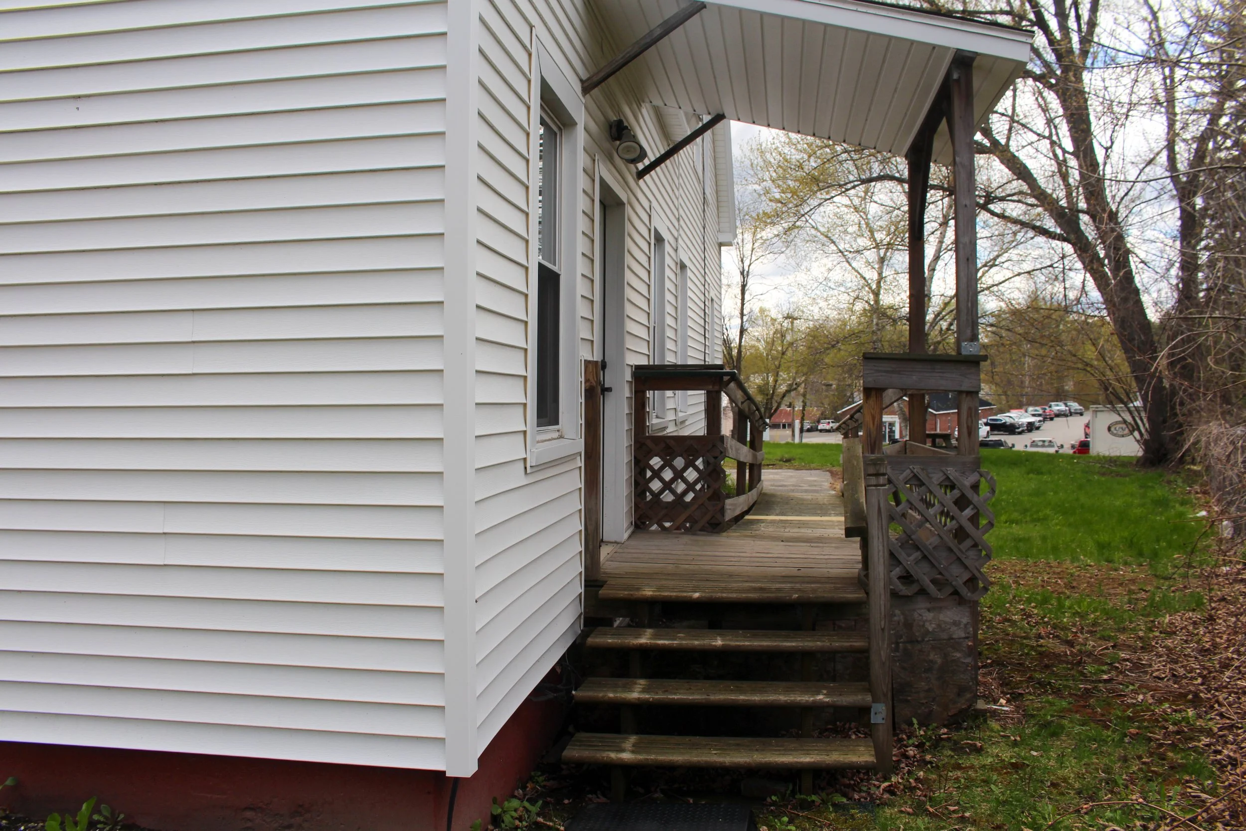 Exterior view of a house with white vinyl siding, small wooden porch, and stairs leading to the yard, with parked cars and trees in the background.