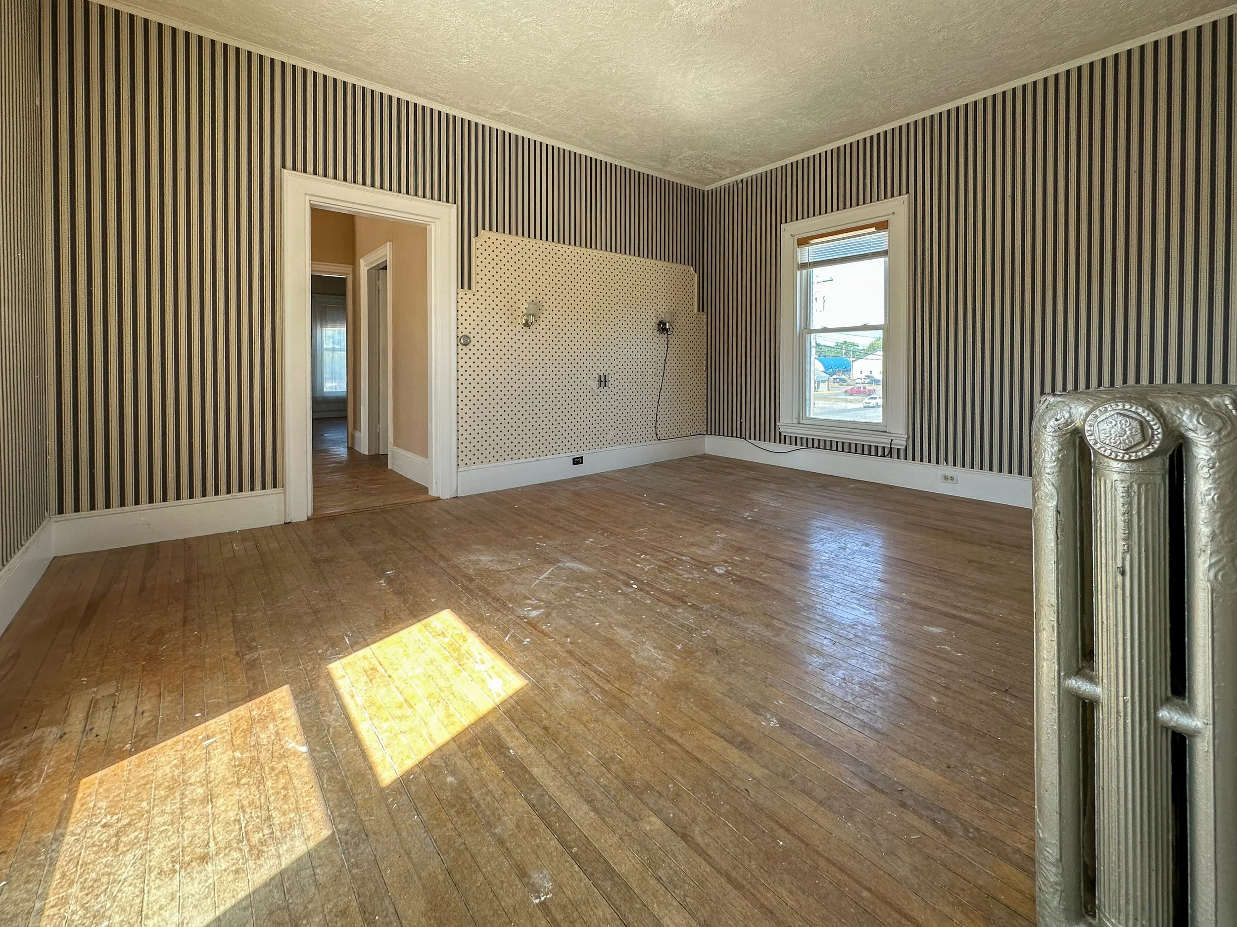 Empty room with wood floor, striped wallpaper, two windows, and a decorative silver fireplace