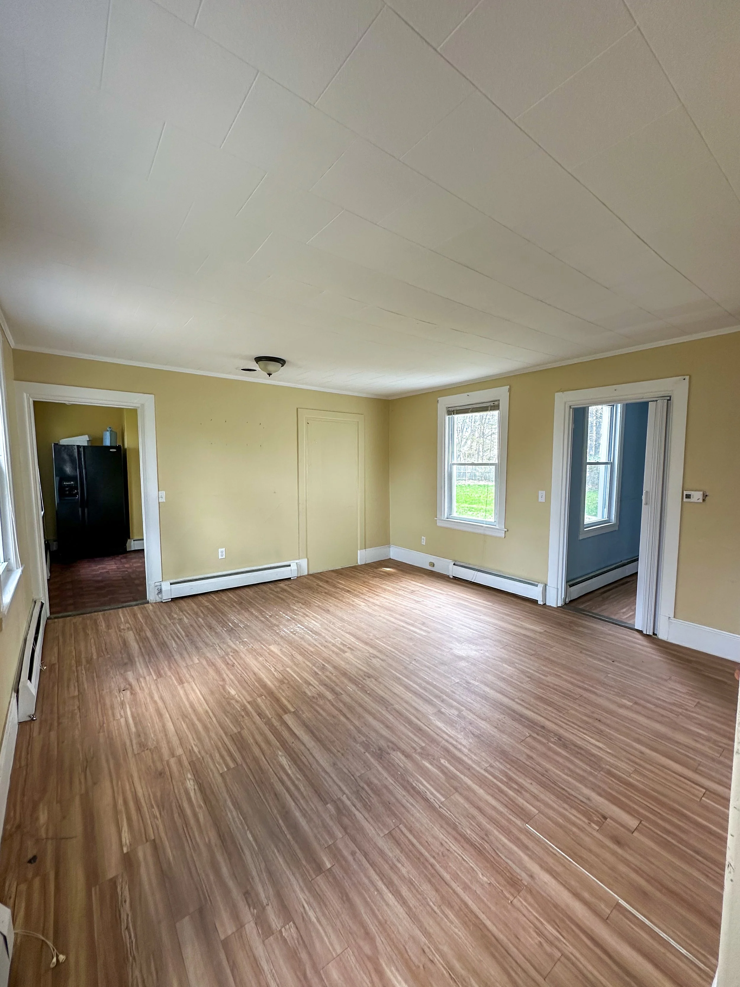 Empty living room with wood flooring, yellow walls, two windows, and a black refrigerator visible in an adjacent room.