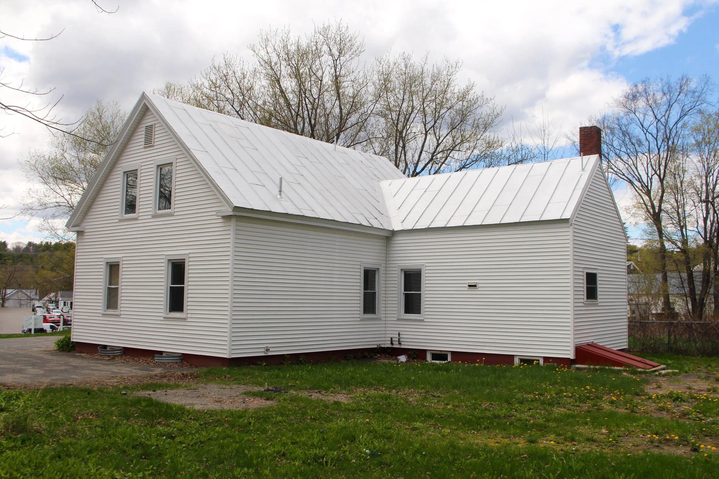 A white house with vinyl siding, a steeply pitched metal roof, and a brick chimney. The house has multiple windows and is surrounded by trees with some with budding leaves. The sky above is partly cloudy.
