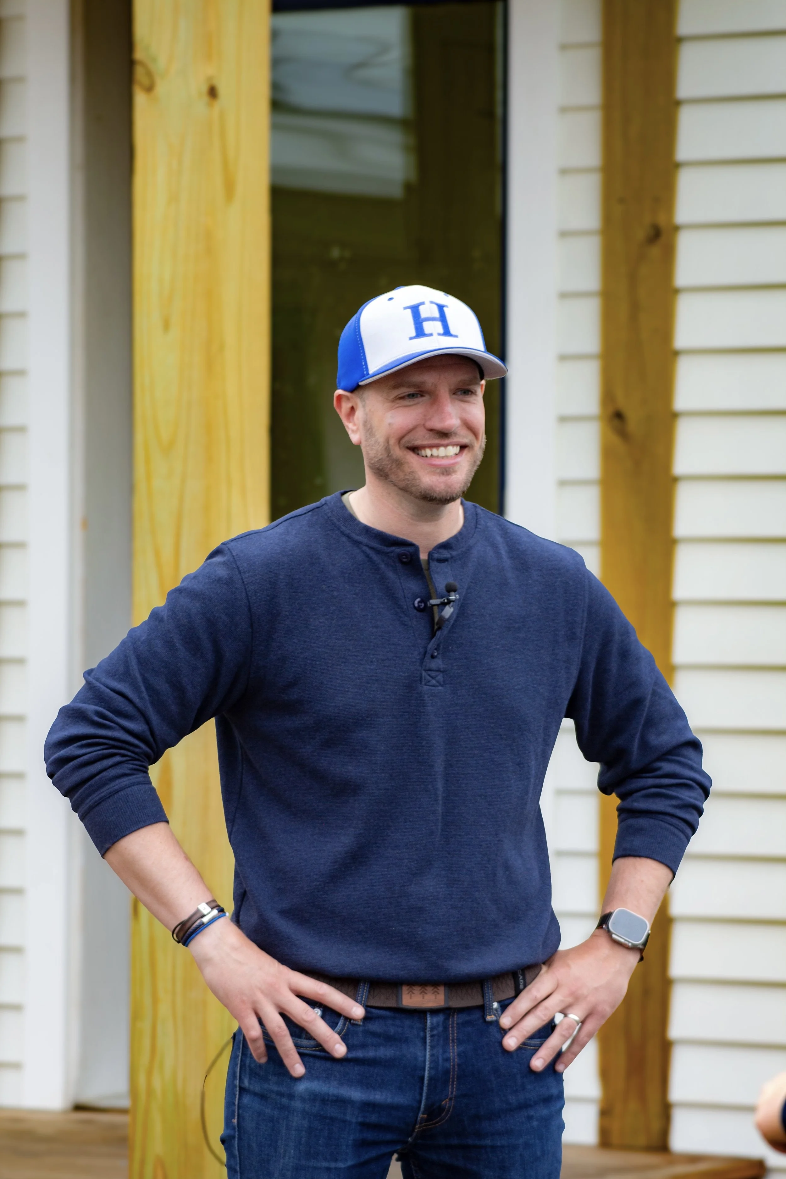 A man wearing a blue and white baseball cap with an 'H' on it, a navy blue long-sleeve shirt, and a smartwatch, standing outdoors in front of a house with beige siding and wooden columns, smiling with his hands on his hips.