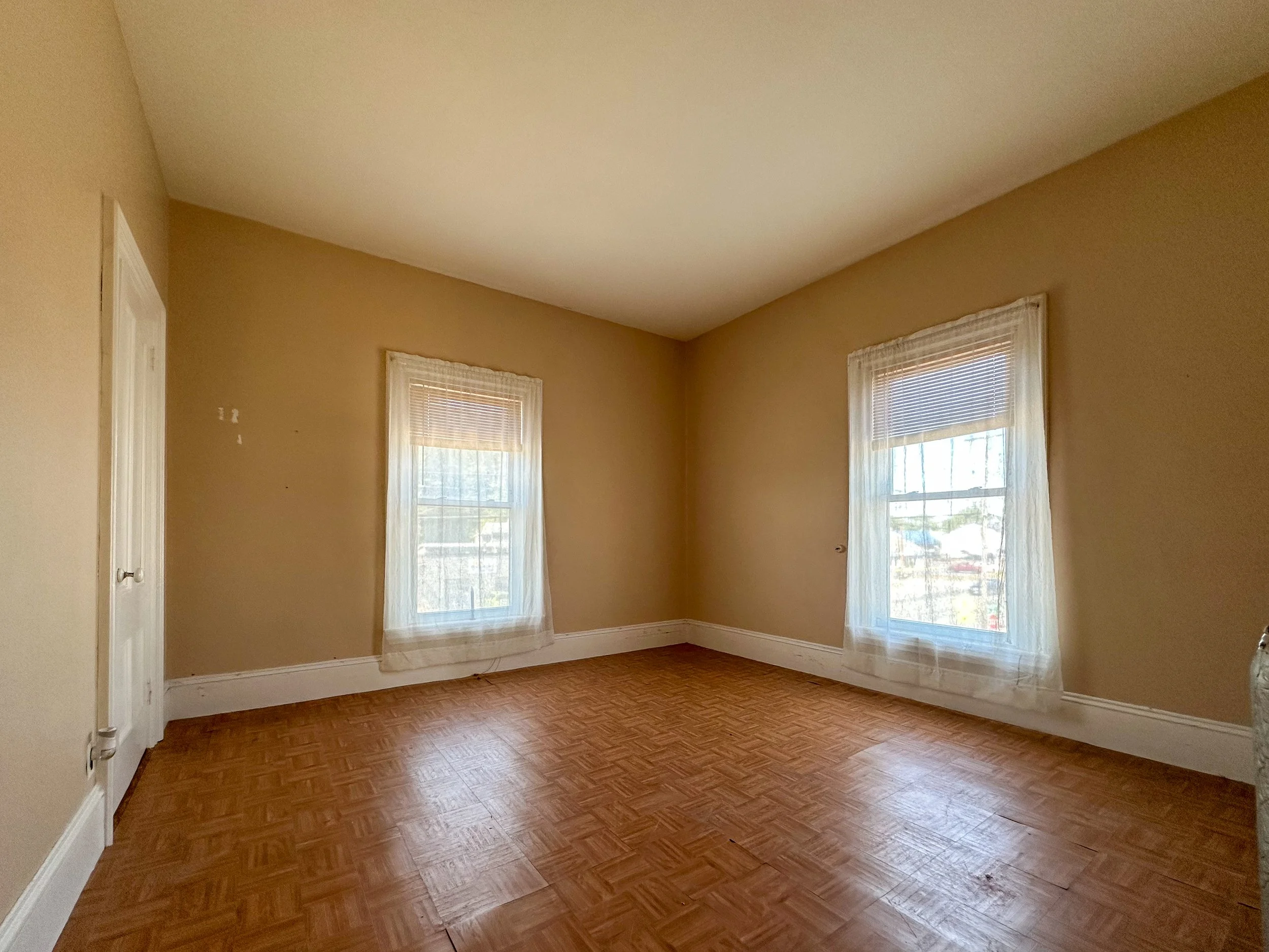 Empty room with beige walls, two windows with blinds, and hardwood flooring.