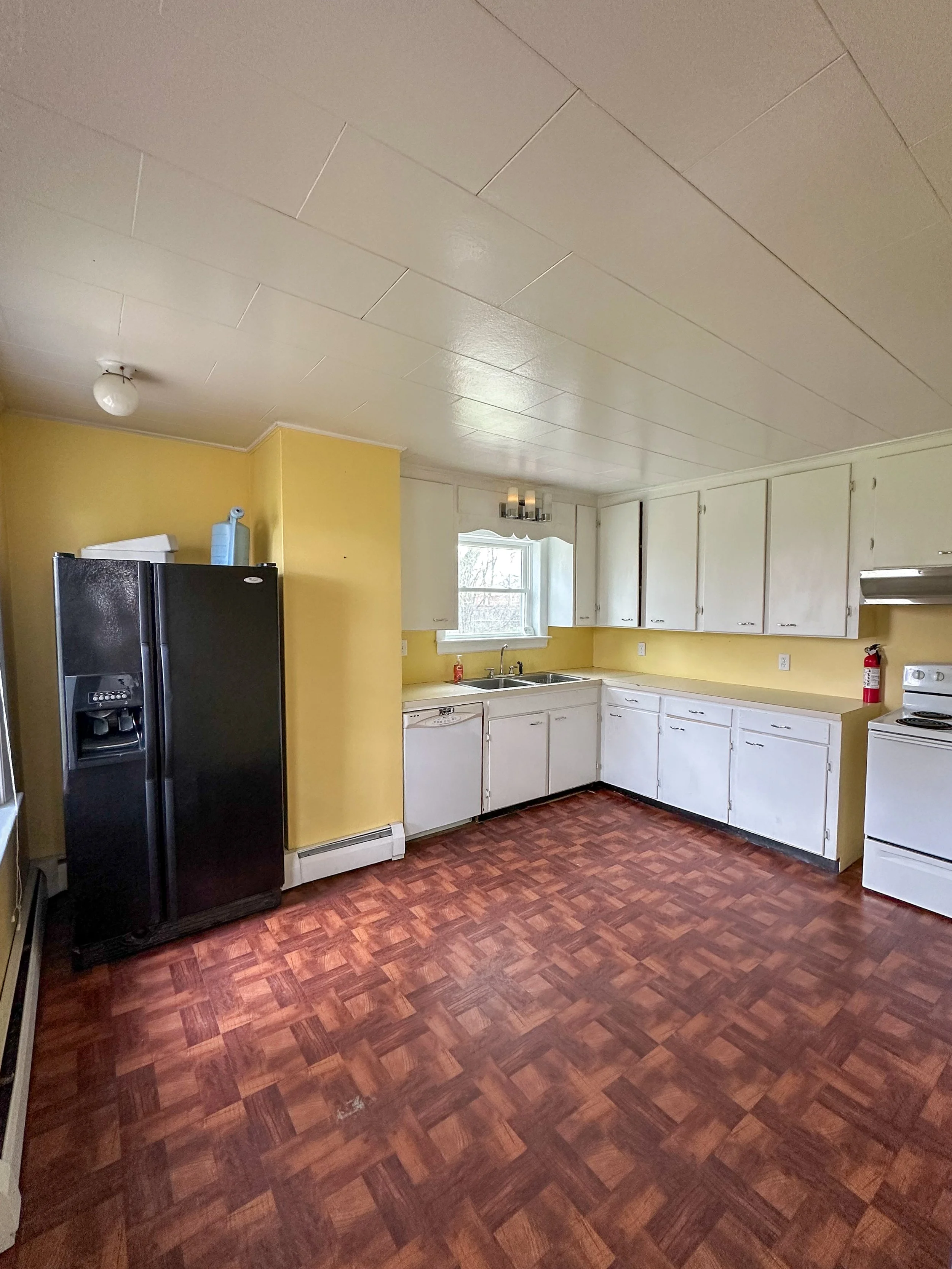 A kitchen with white cabinets, a yellow wall, and a brown patterned floor. There is a black refrigerator, a window above the sink, and a fire extinguisher on the right side. The ceiling has a white finish with a light fixture.