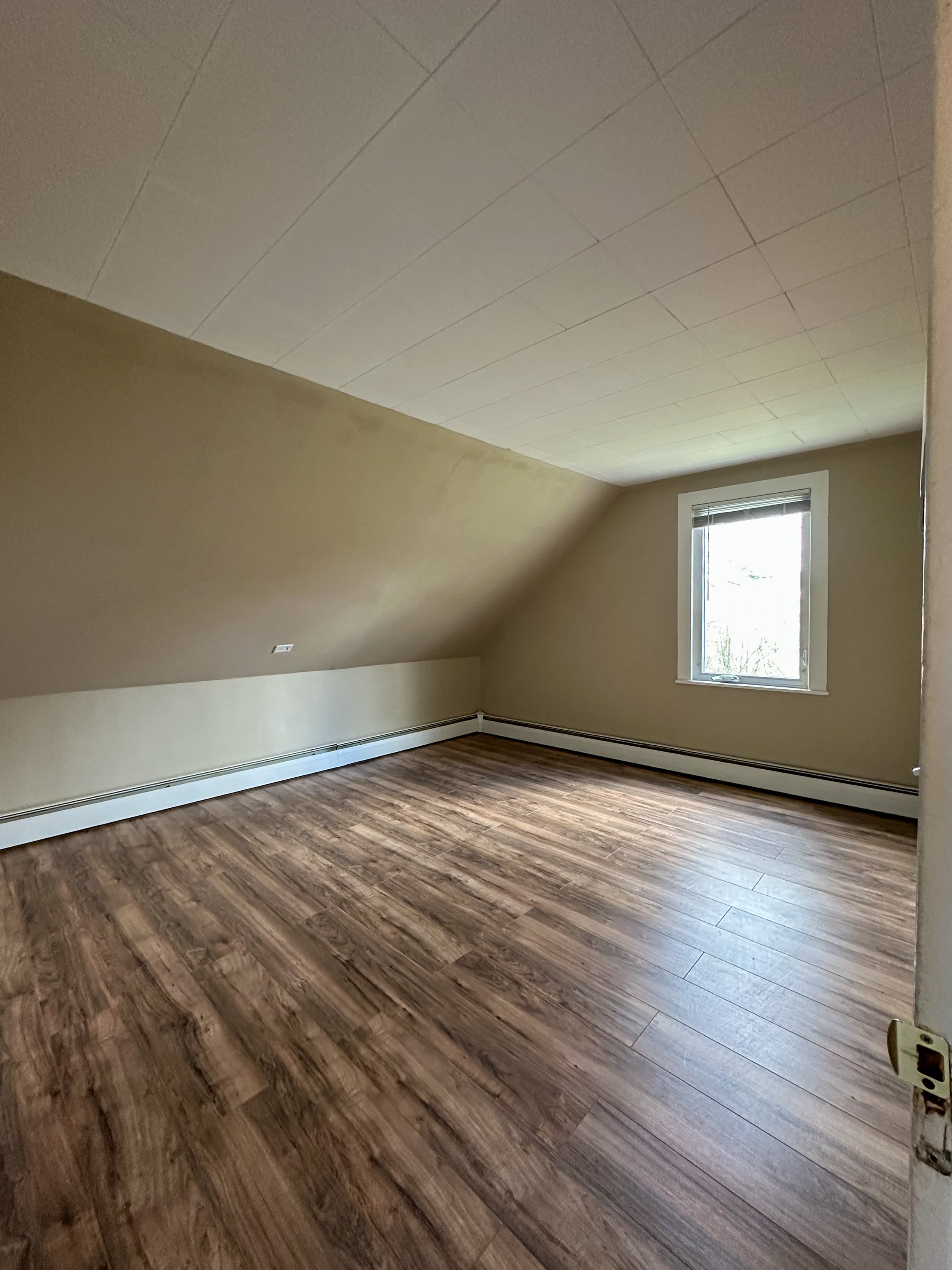 Empty room with wood laminate flooring, beige walls with a sloped ceiling, a window on the far wall, and an electrical outlet near the floor.