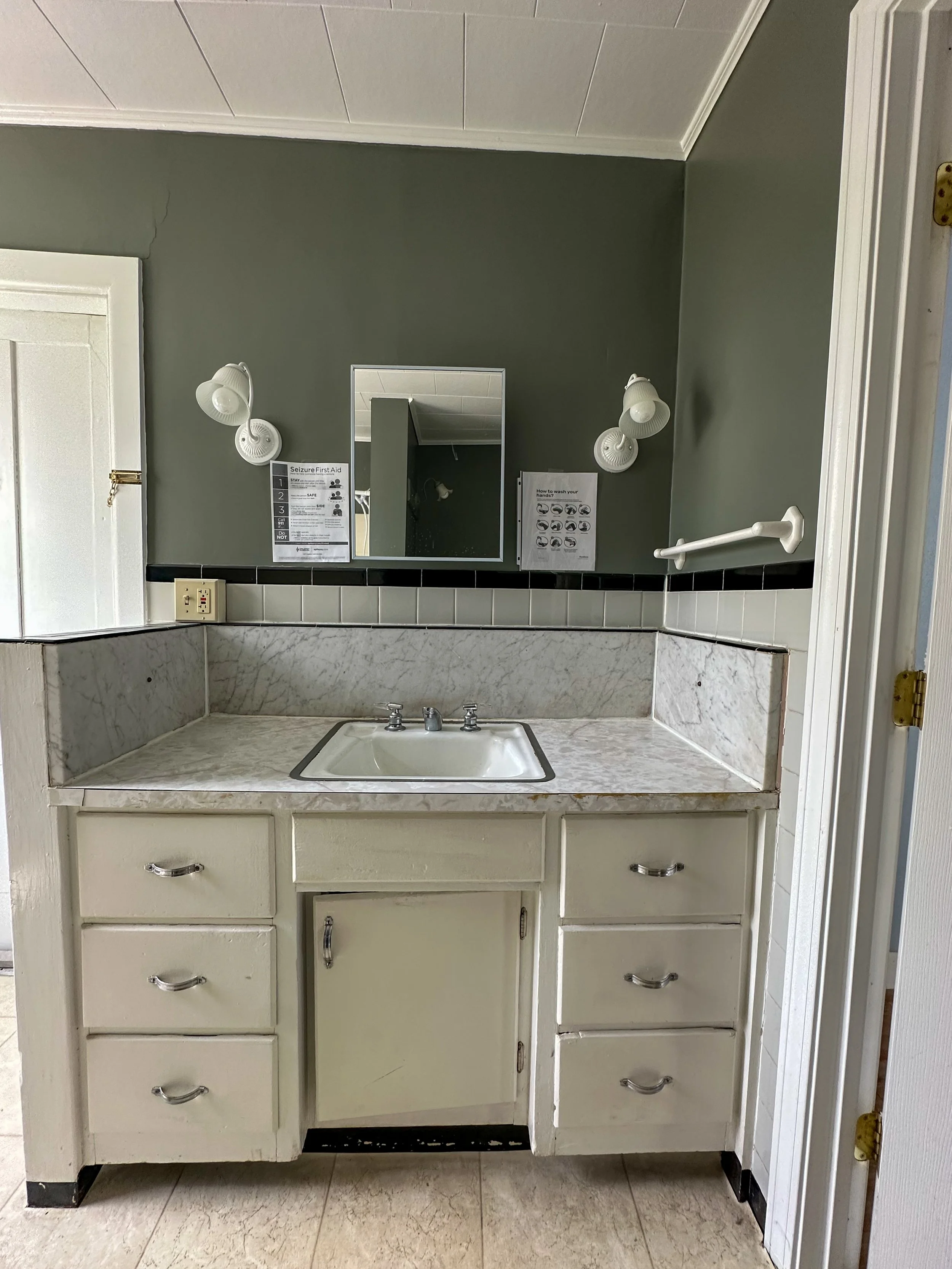 Bathroom mirror above a sink, with two white wall-mounted lights, a sign about seizures and handwashing instructions, and a white cabinet with drawers below the sink.