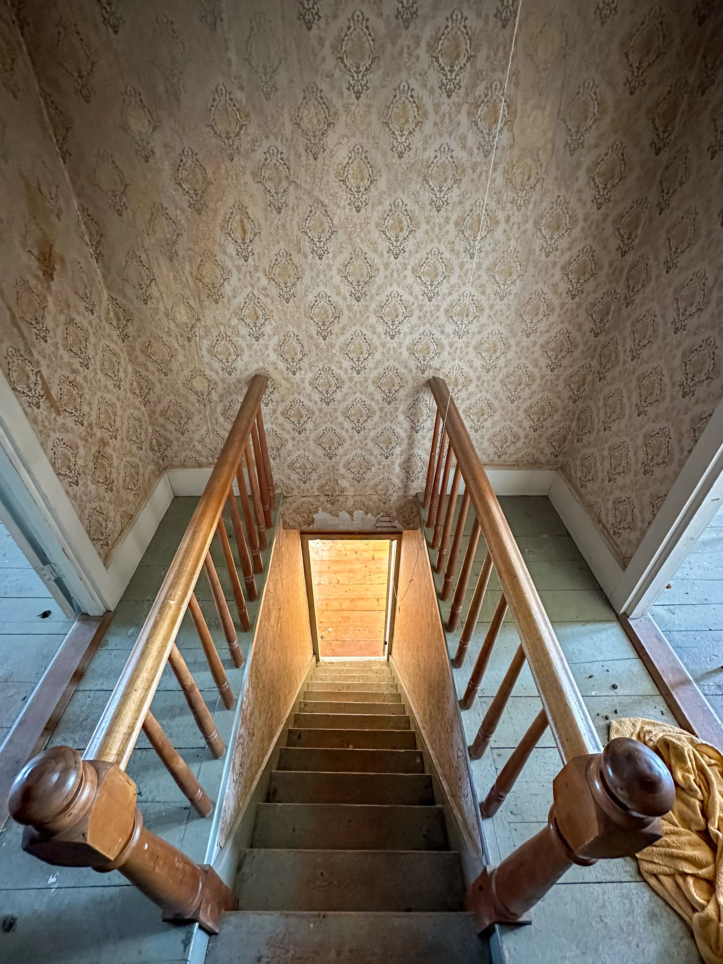 From the top of a staircase looking down, wooden handrails on both sides, wallpaper with a light pattern on the walls, tiled floor, and a partially visible shoe or boot at the bottom right.