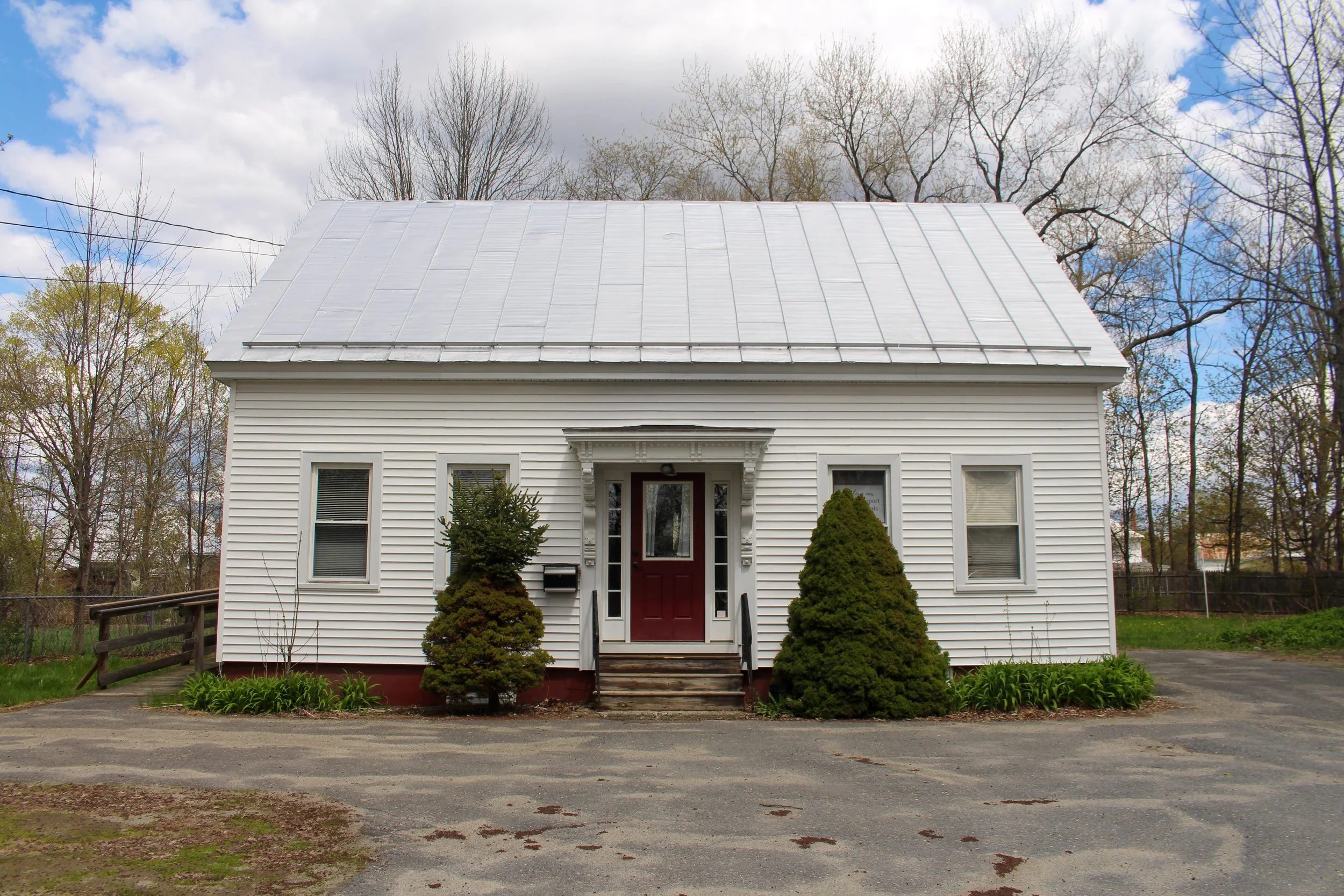 White house with metal roof, surrounded by bushes and trees, with cloudy sky.