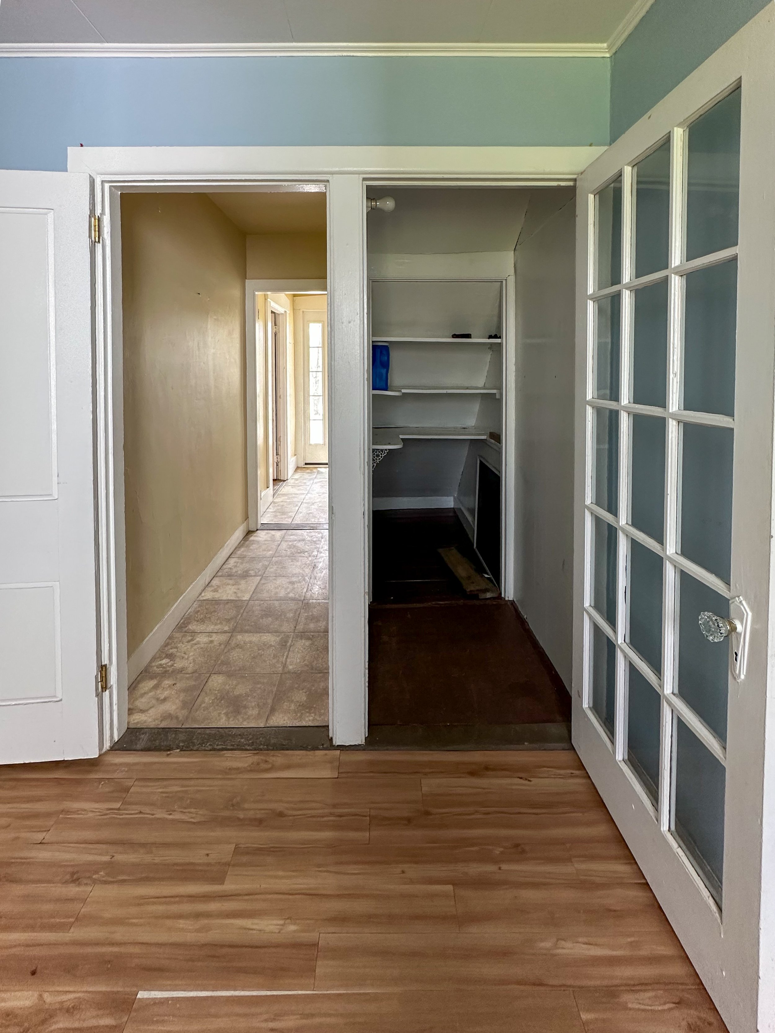 Image of an interior view showing a doorway leading to a hallway, with a small room with white shelves on the right, and a floor transition from wood to tile.