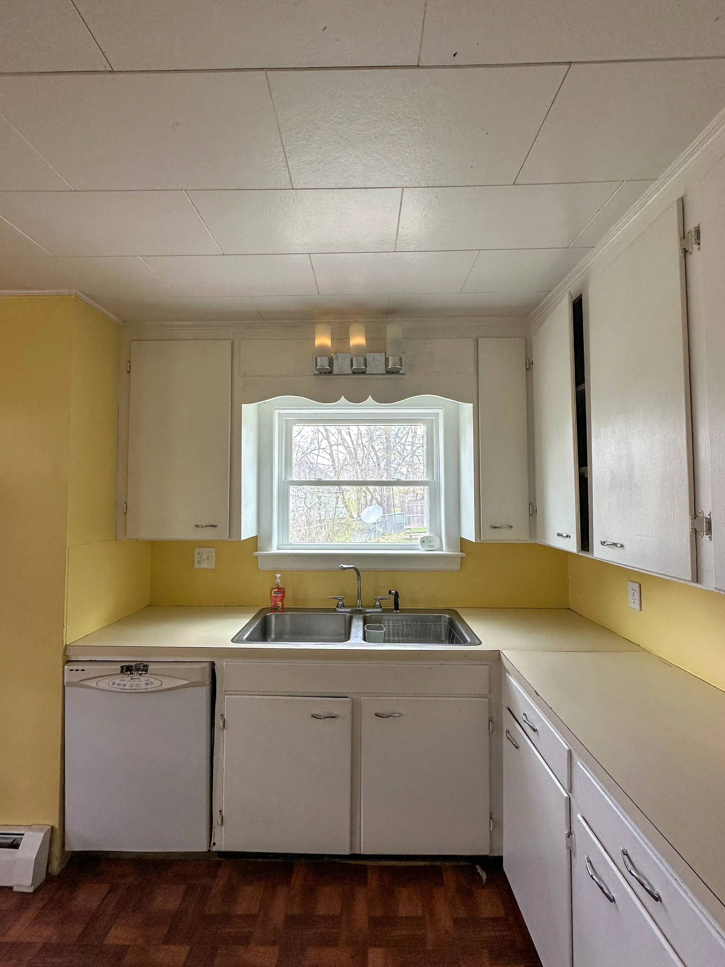 Kitchen with white cabinets, a window above the sink, and a yellow wall. There is a sink with a faucet, soap dispenser, and a small white cup. The floor is wood or laminate. The ceiling has white tiles and a light fixture with three bulbs.