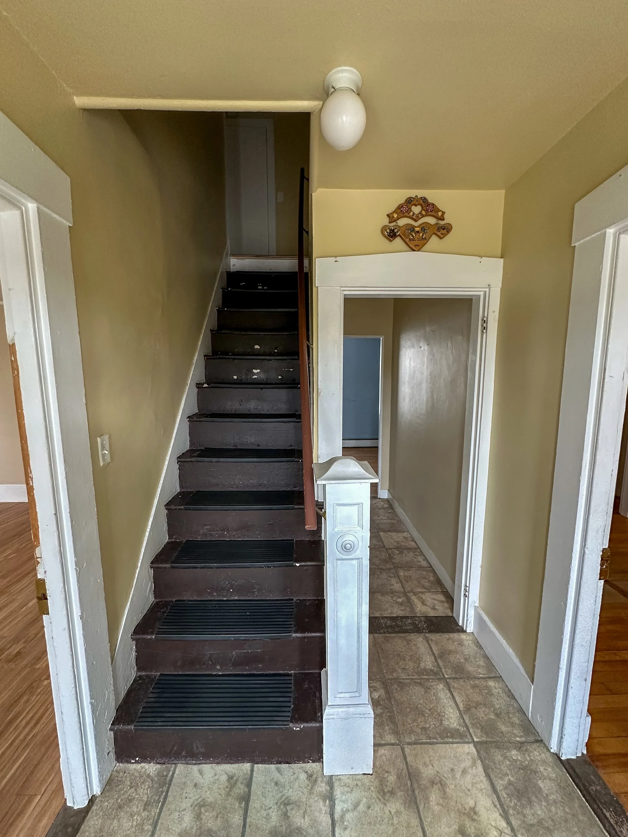 Interior view of a staircase with black steps, a short white wooden railing, beige walls, a hallway with tiled floor, and a doorway leading to another room. There is a decorative wall hanging with hearts and the word 'Welcome' above the door.