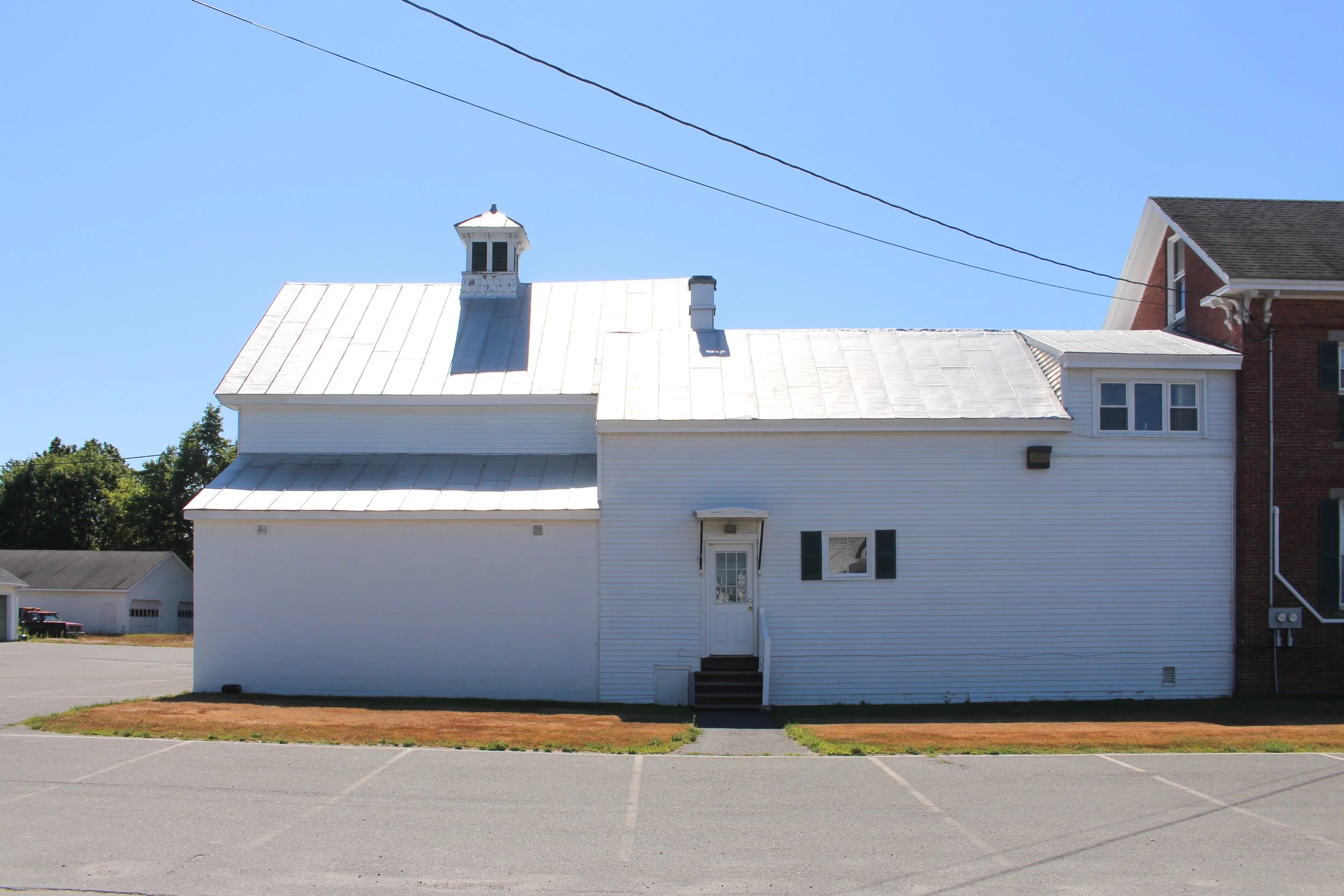 A white residential building with a metal roof, a small elevated porch with stairs, and a window on the second floor. The building is located in a parking lot with marked spaces, and there are trees and other houses in the background under a clear bl