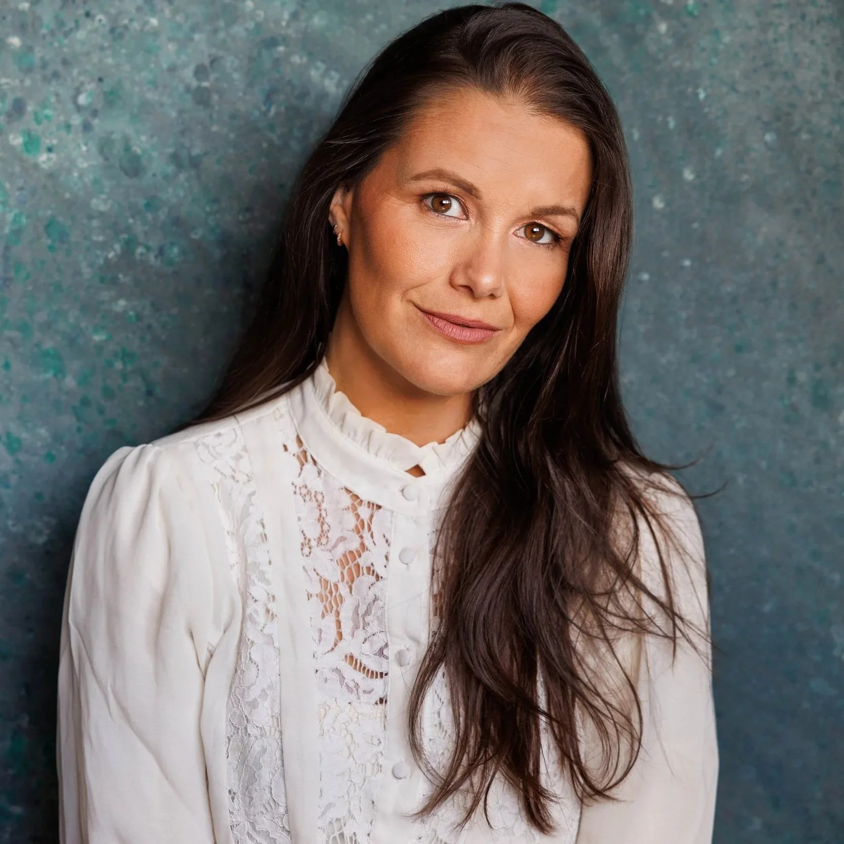 A woman with long brown hair and brown eyes wearing a white lace blouse with a ruffled collar, posing against a speckled teal background.
