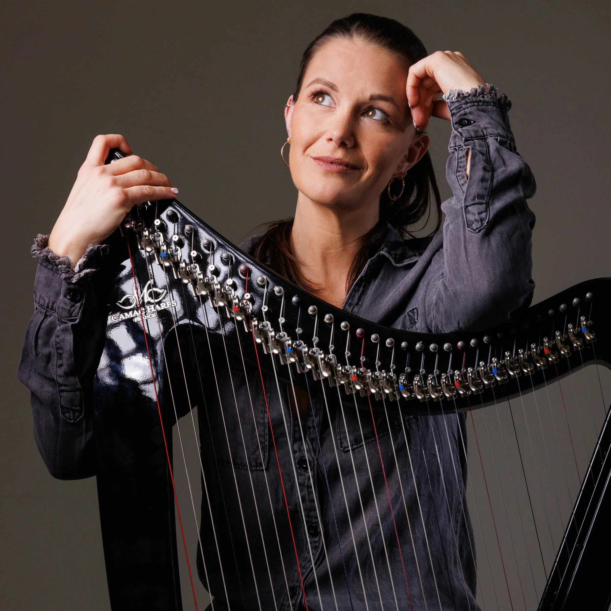 A woman with dark hair and brown eyes looking thoughtfully while holding a round object, standing next to a harp with colorful strings.