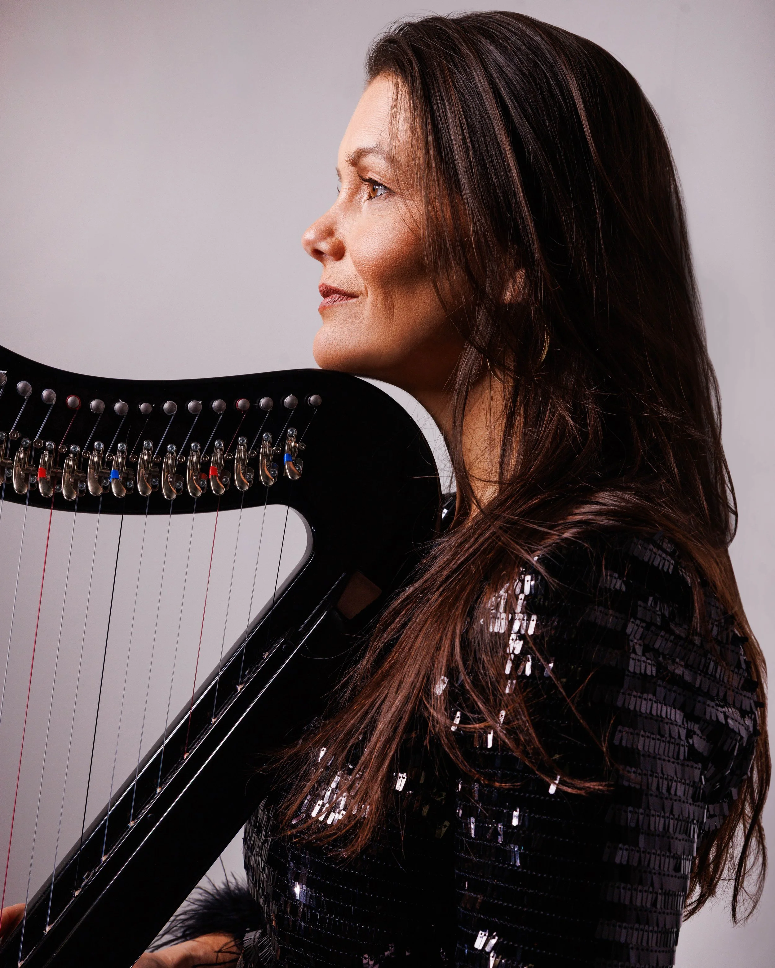A woman with long brown hair and light makeup posing with a harpsichord against a plain gray background.