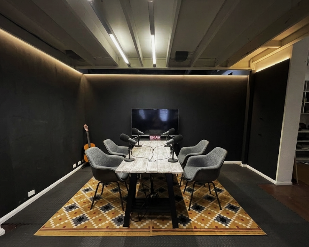 A podcast recording studio with a table, six gray chairs, six microphones, a TV screen, and a guitar in the corner, with a patterned rug and black walls.