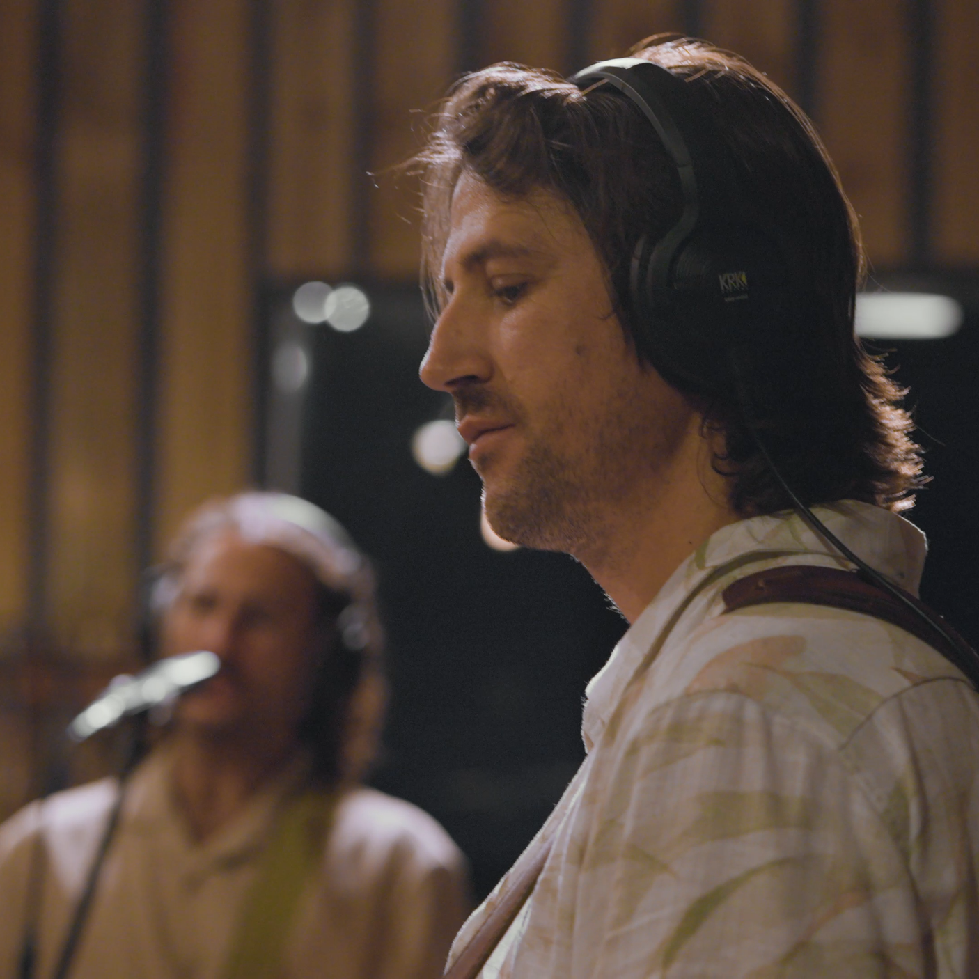 A man with long dark hair and fair skin wearing a plaid shirt and headphones, standing in a recording studio with a woman in the background also wearing headphones.