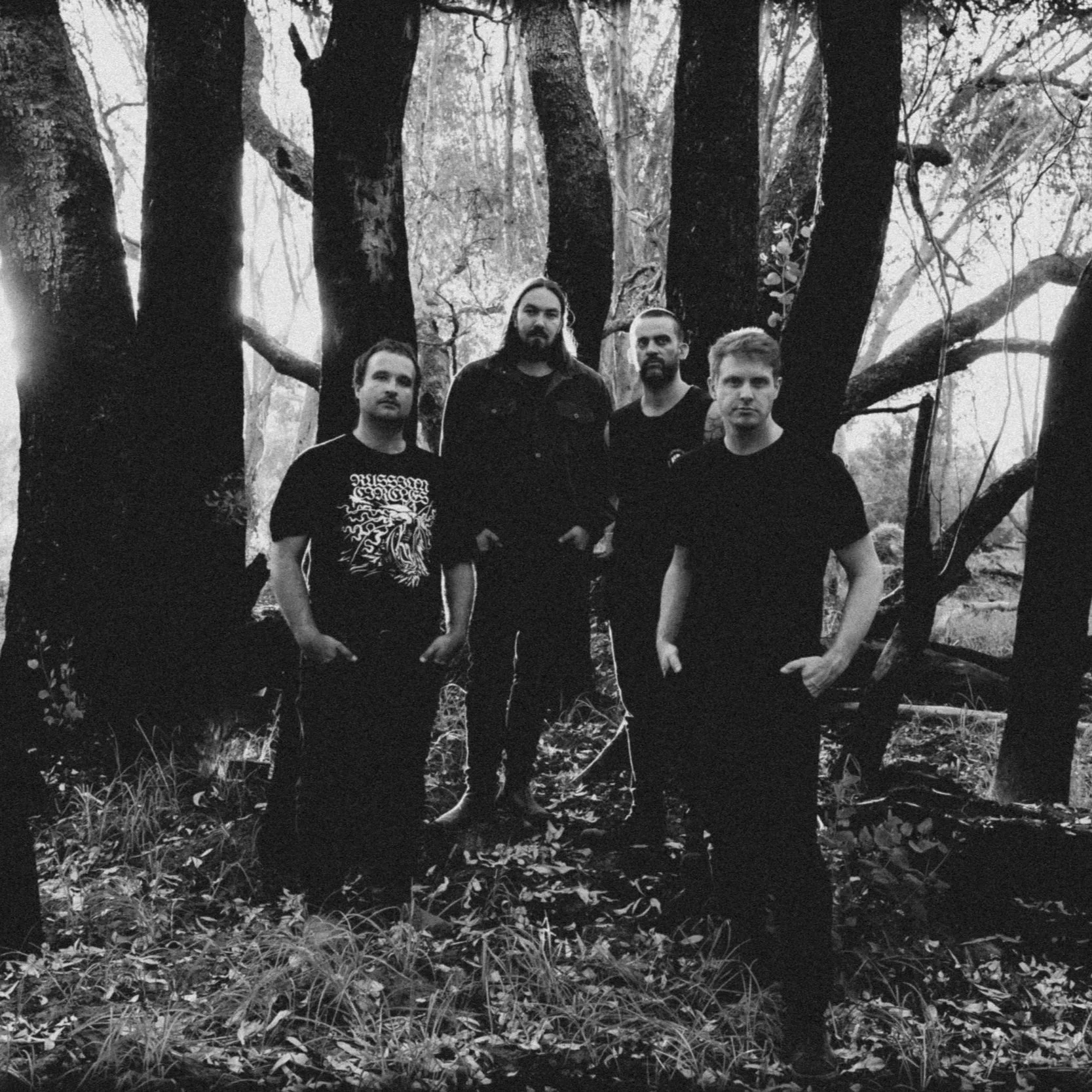 Four men standing in a wooded area with trees and foliage in black and white.