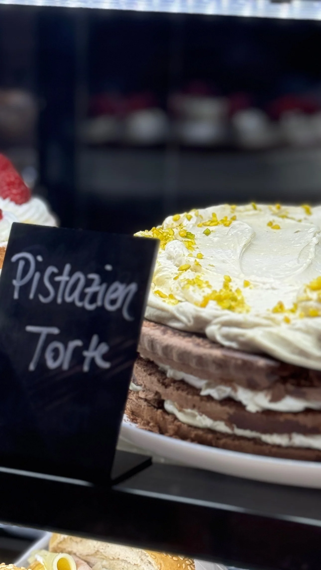 Kuchen mit weißer Creme, bestreut mit gelben Schuppen, auf einem Tortenteller. Im Vordergrund eine Tafel mit der Aufschrift 'Pistazien Torte'.